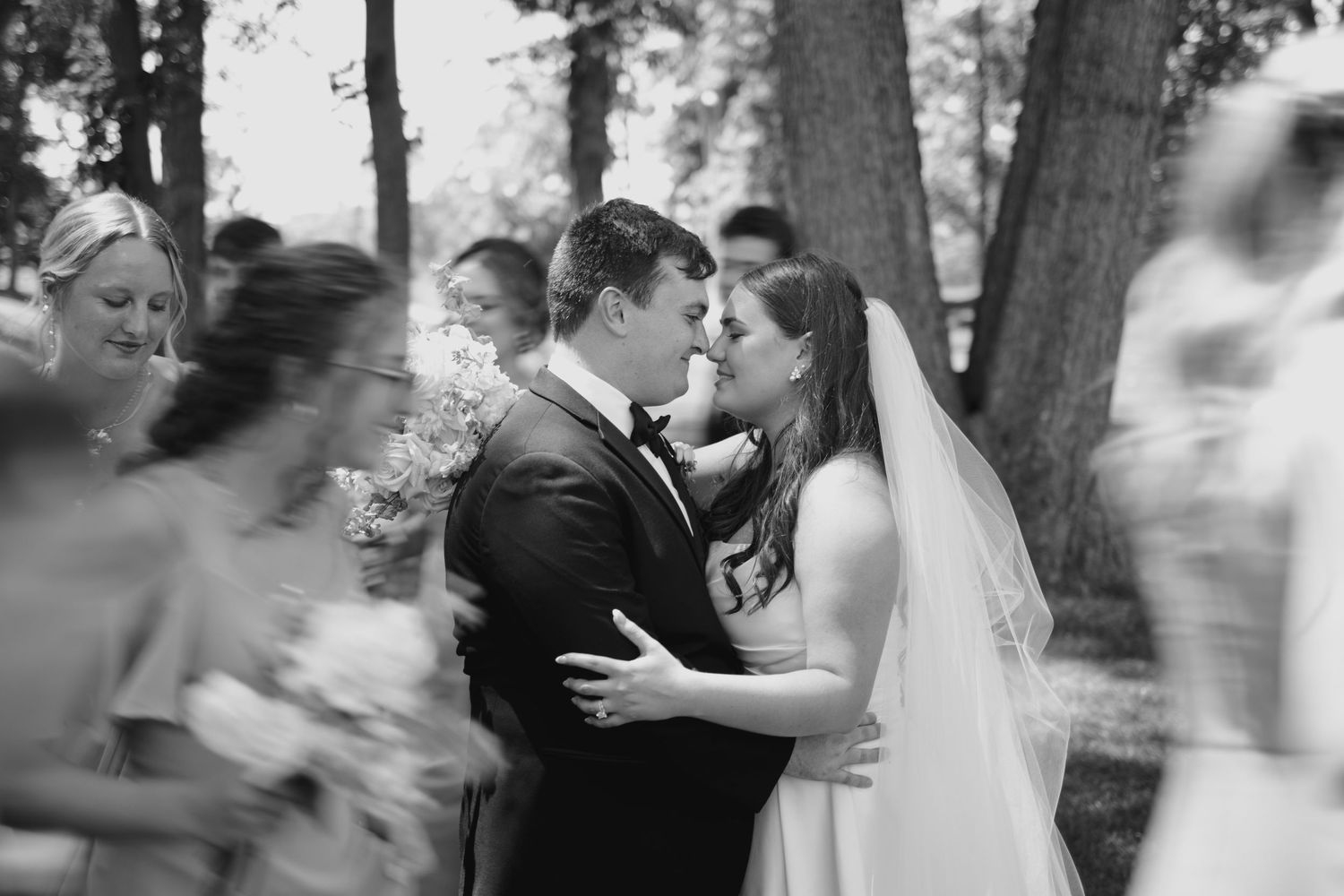 Black and white photo of a bride and groom sharing a kiss surrounded by blurred wedding party outdoors.