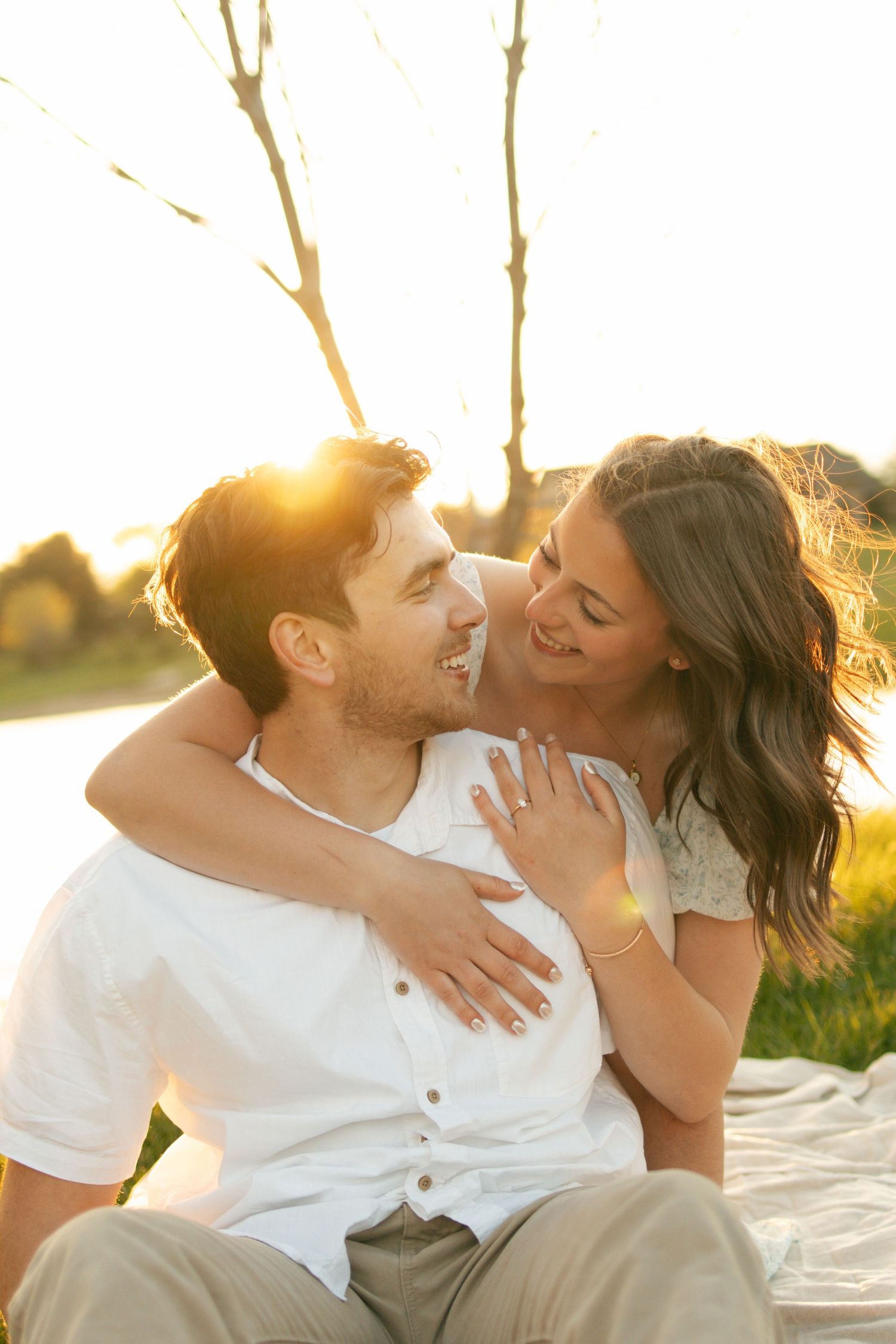 Happy couple sharing a romantic moment outdoors at golden hour sunset, woman hugging man from behind while laughing.