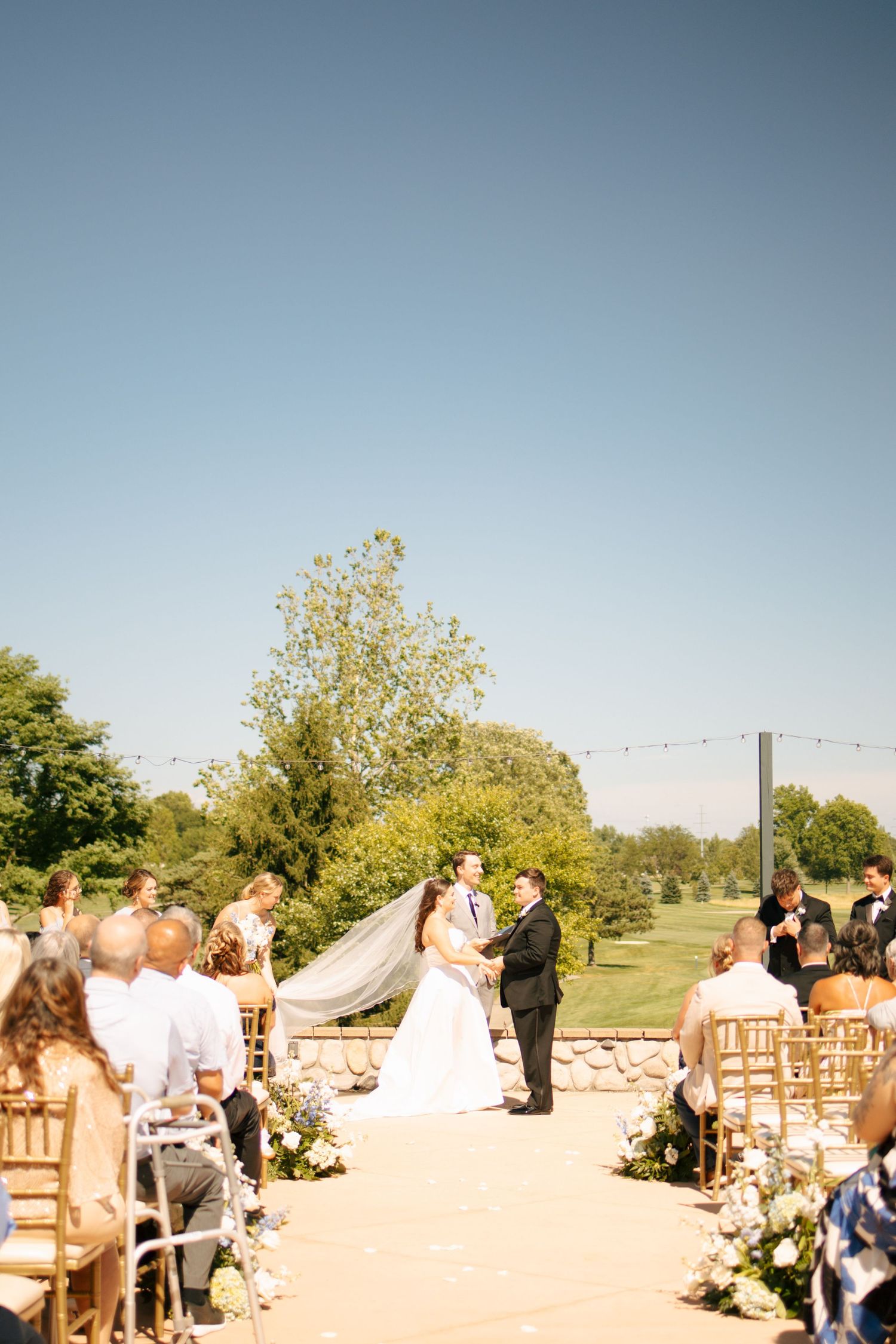 Outdoor wedding ceremony with bride and groom at the altar, guests seated in gold chairs, lush greenery and clear blue sky.