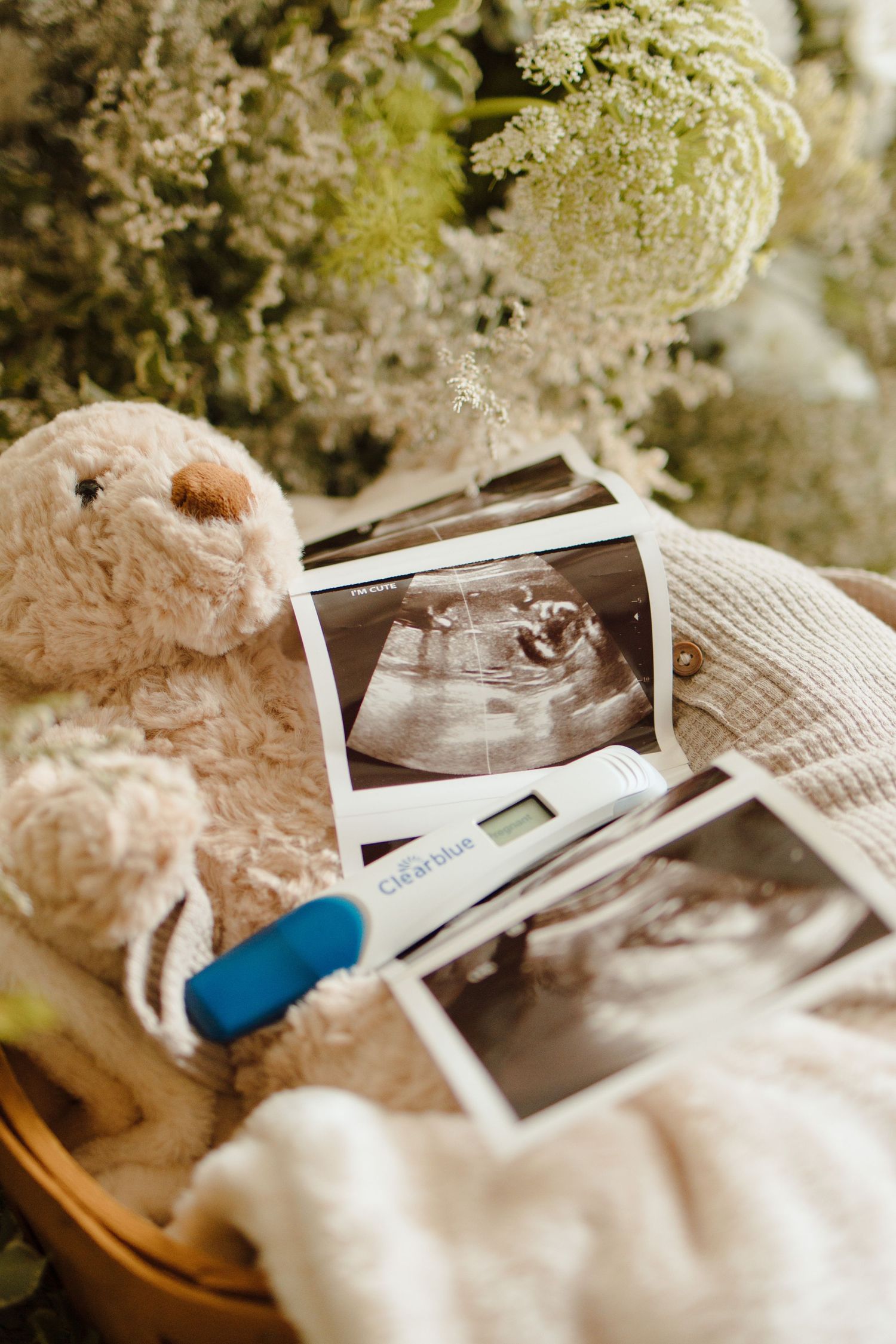 Pregnancy announcement flat lay with ultrasound photos, Clearblue test, teddy bear, white knit blanket, and flowers.