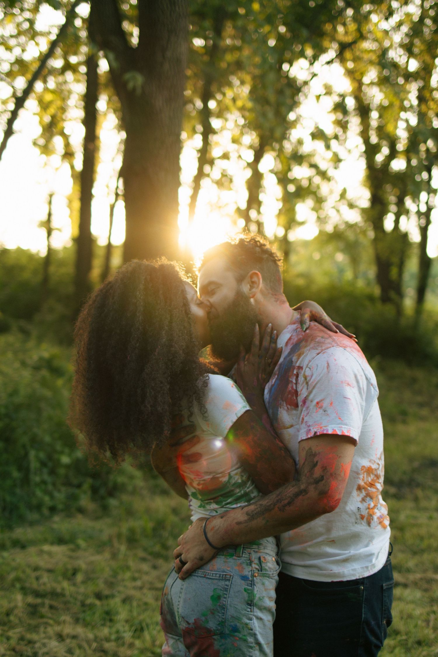 Couple sharing a romantic kiss at golden hour in a lush forest, both wearing paint-splattered white shirts.