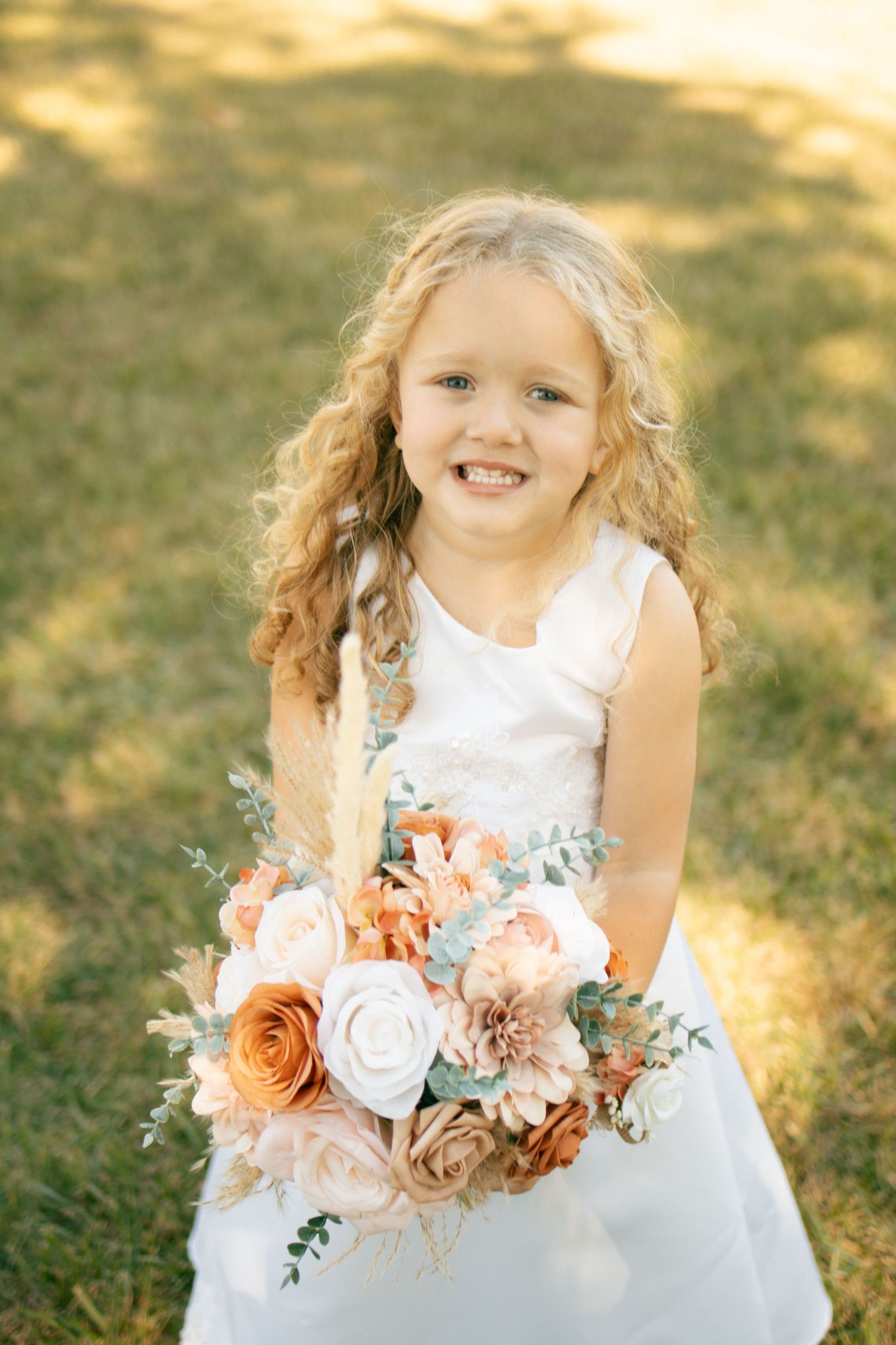 Smiling daughter of bride and groom in white dress holding peach and blush floral bouquet on sunny grass lawn.