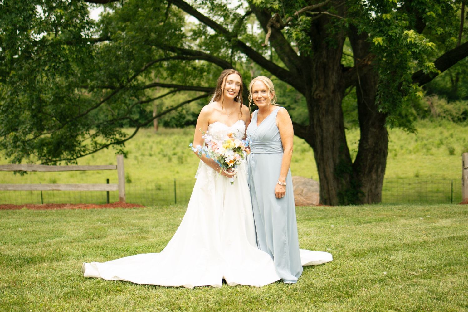 Bride in white gown holding bouquet stands with bridesmaid in light blue dress on green lawn near wooden fence.