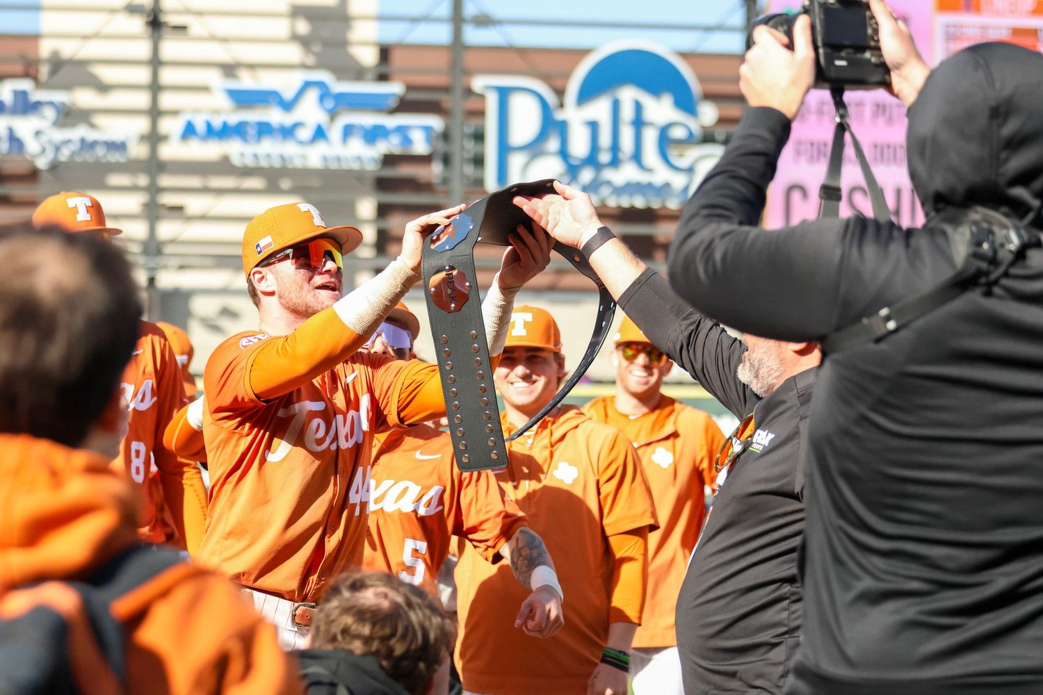 Baseball team members in bright orange jerseys raise their hands in celebration during a game while being photographed.