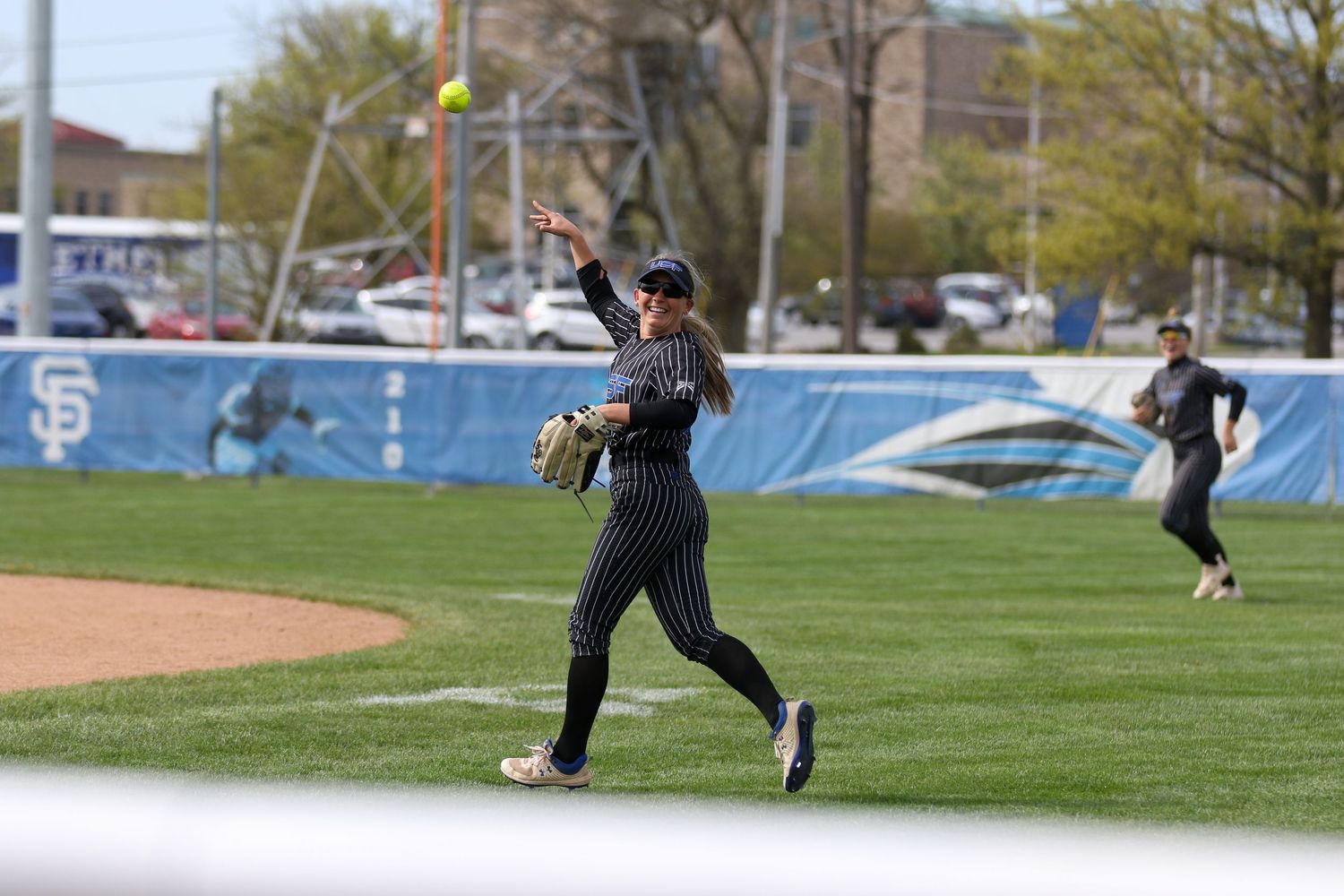 Baseball player in navy pinstriped uniform throws the ball during a sunny game on grassy field with blue fence in background.