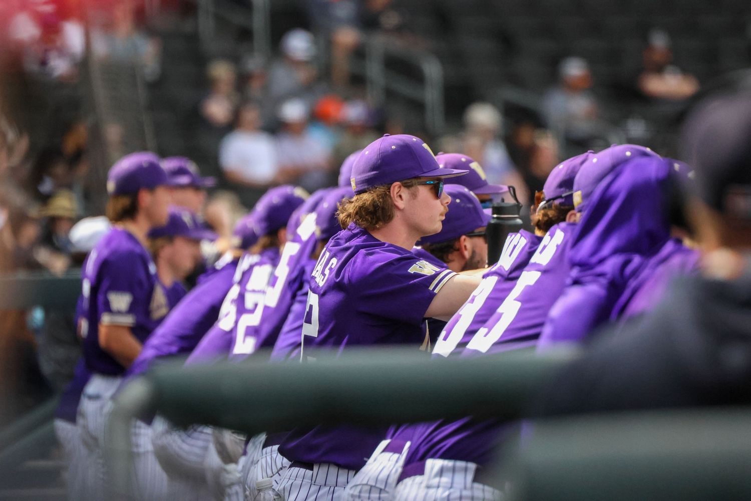 Baseball players in purple uniforms gather in the dugout during a game while spectators watch from the stands.