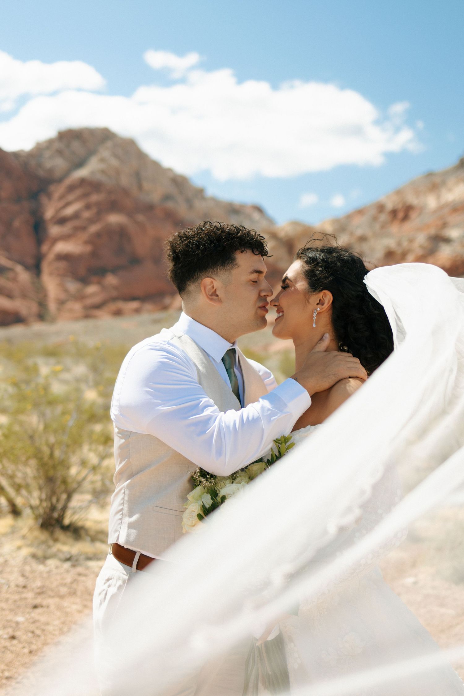 A newlywed couple shares an intimate moment against a backdrop of red rock formations in the desert.