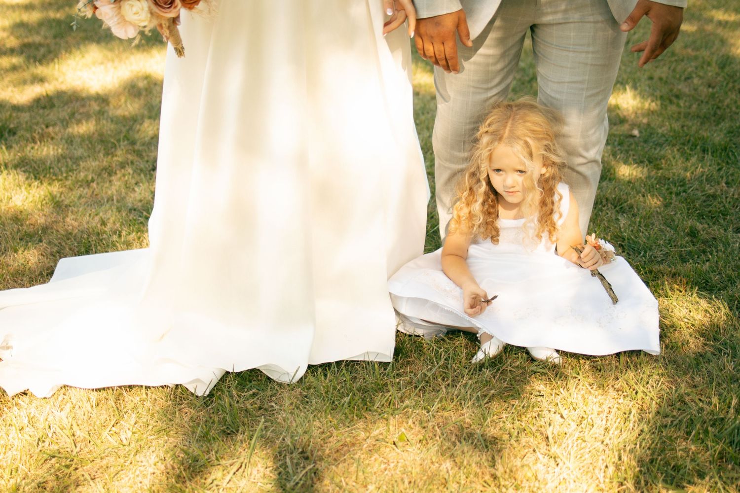 Flower girl in white dress kneeling on grass beside bride's wedding gown train and groom's feet.