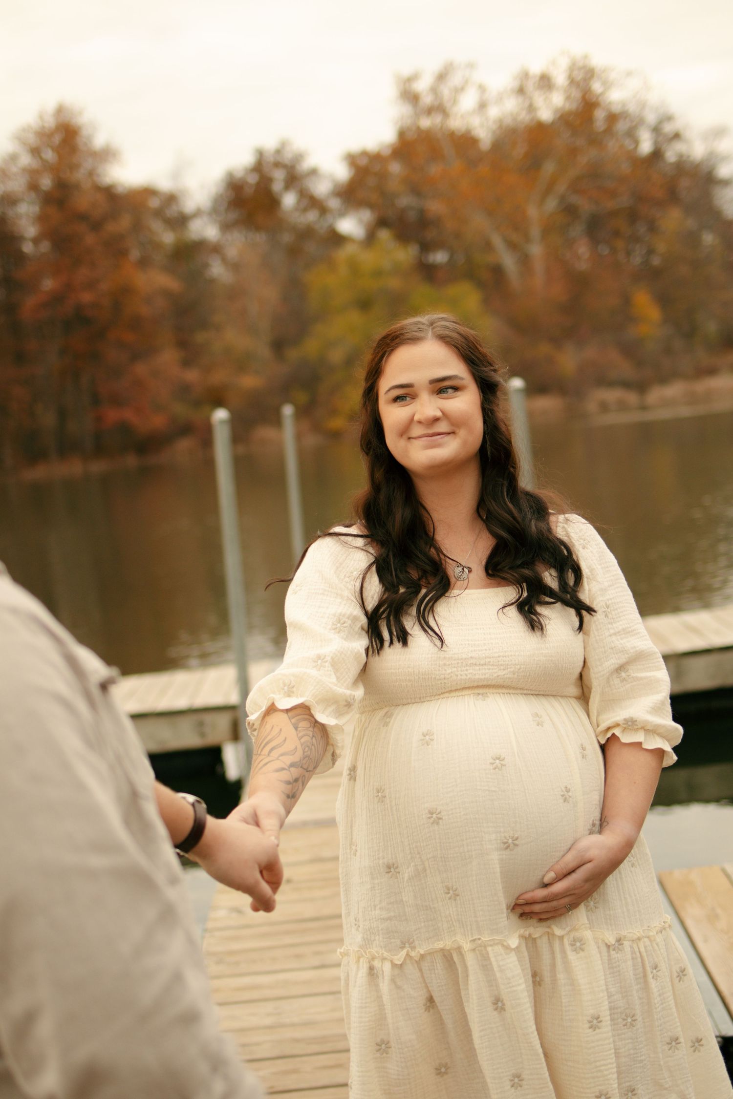 Pregnant woman in cream dress holds hands with partner on autumn lakeside dock, smiling warmly.