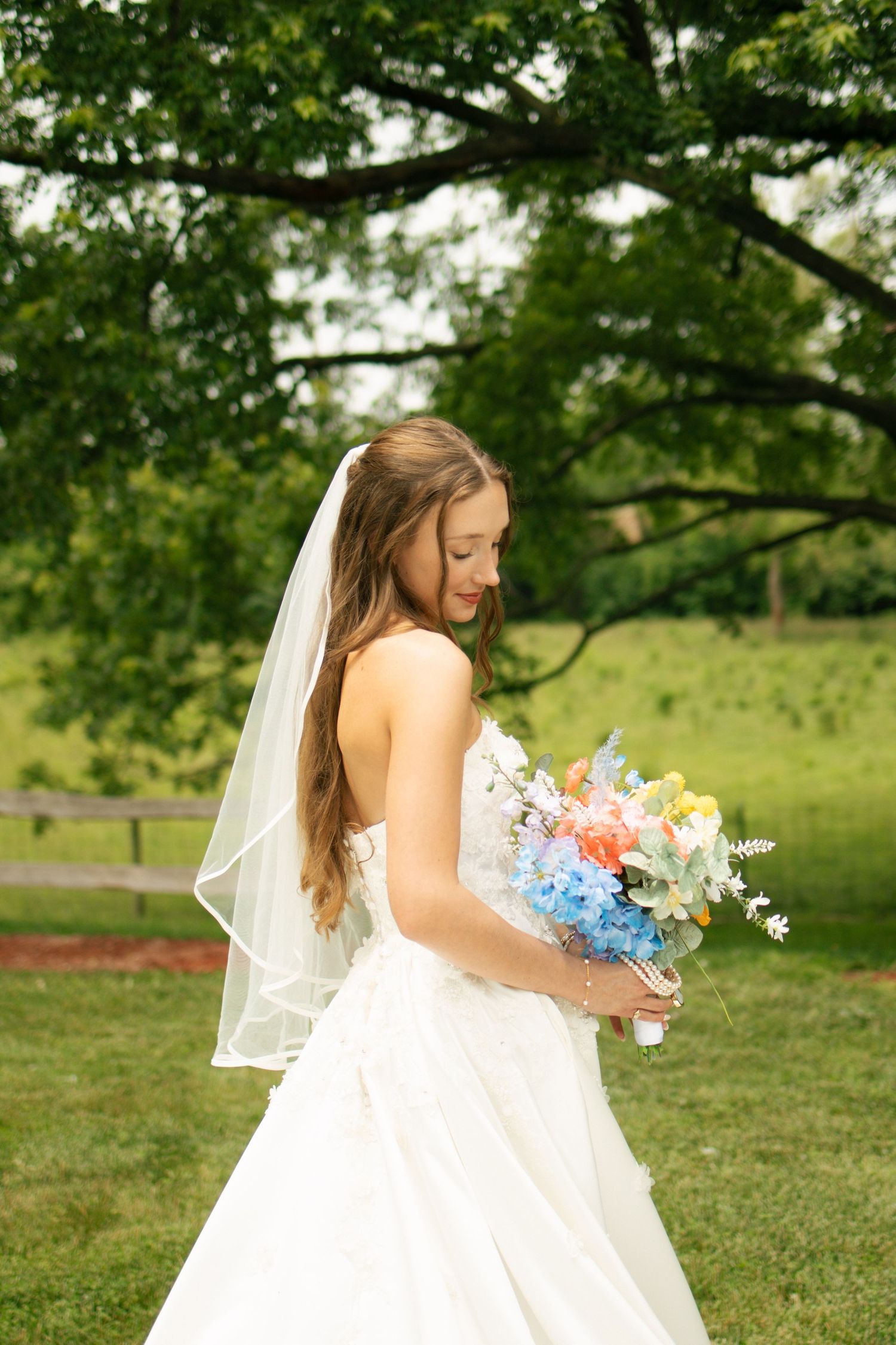 Bride in strapless white gown and veil holds colorful bouquet in lush green outdoor setting.