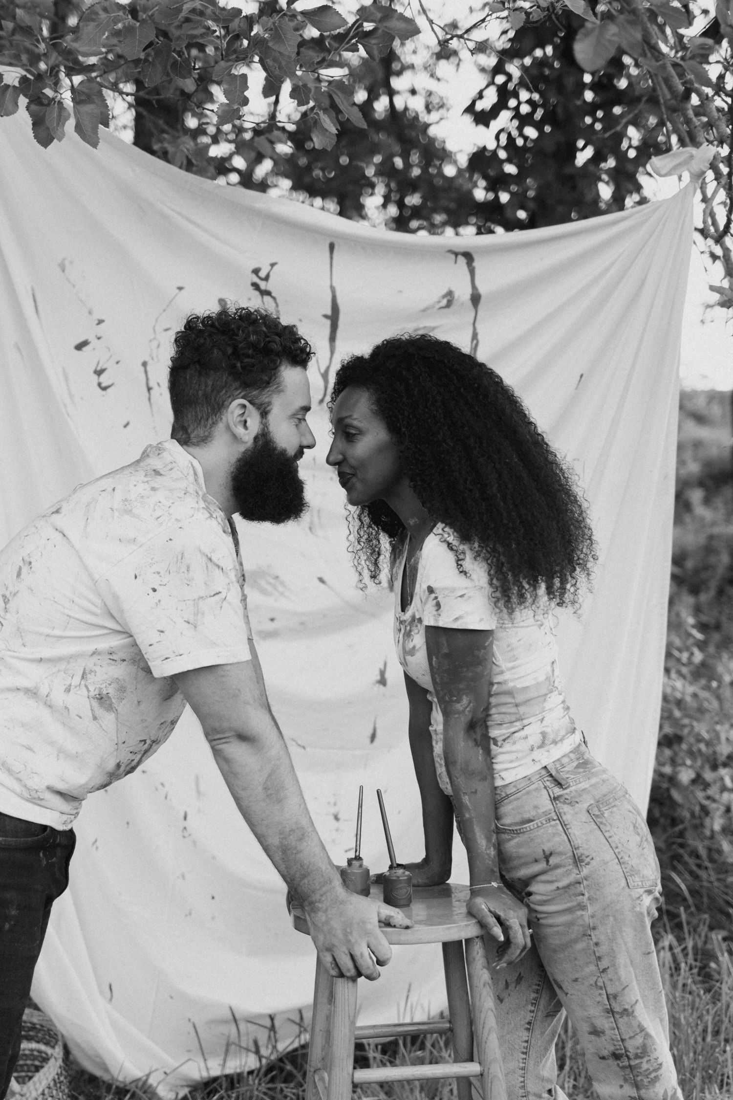 Black and white photo of a bearded man and curly-haired woman in paint-covered clothes sharing a romantic moment outdoors.