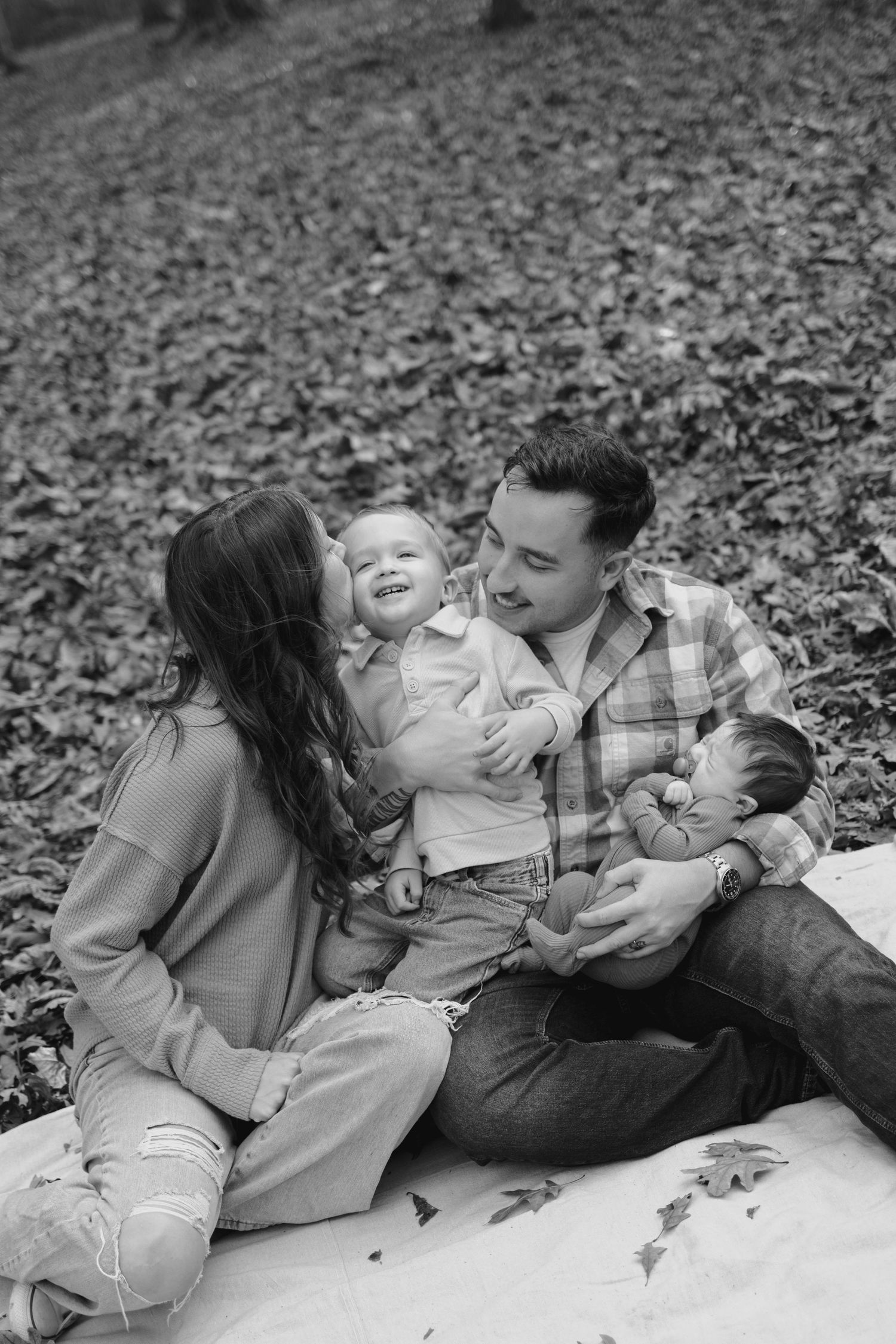 Black and white photo of happy family sitting on blanket in autumn leaves with two young children.