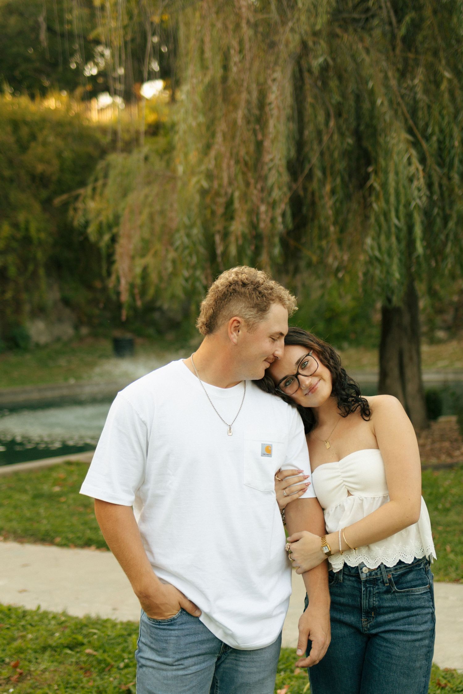 A couple shares a tender moment outdoors near a willow tree, the woman leaning on the man's shoulder.
