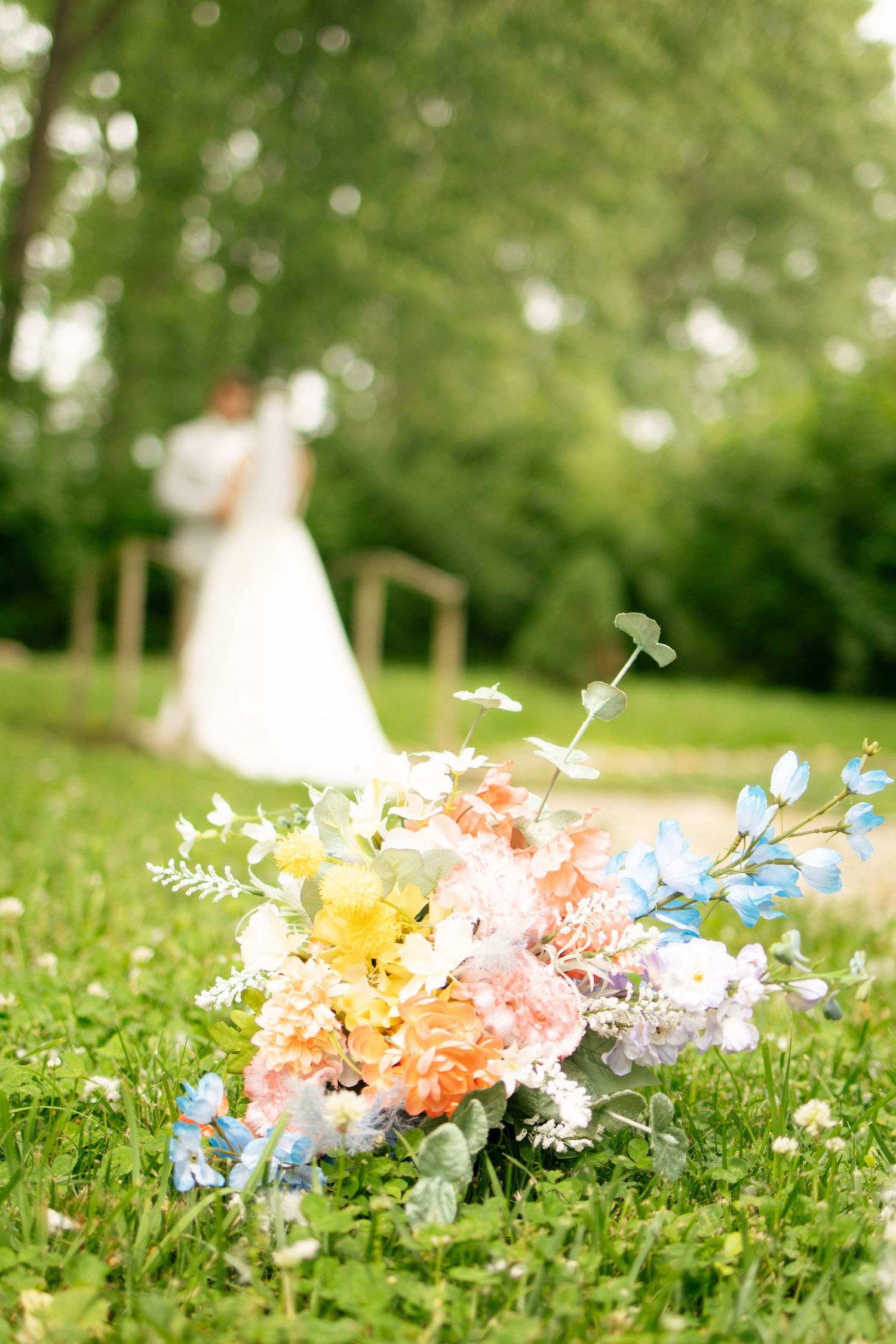 A colorful bridal bouquet rests on green grass with a bride and groom embracing softly in the background.