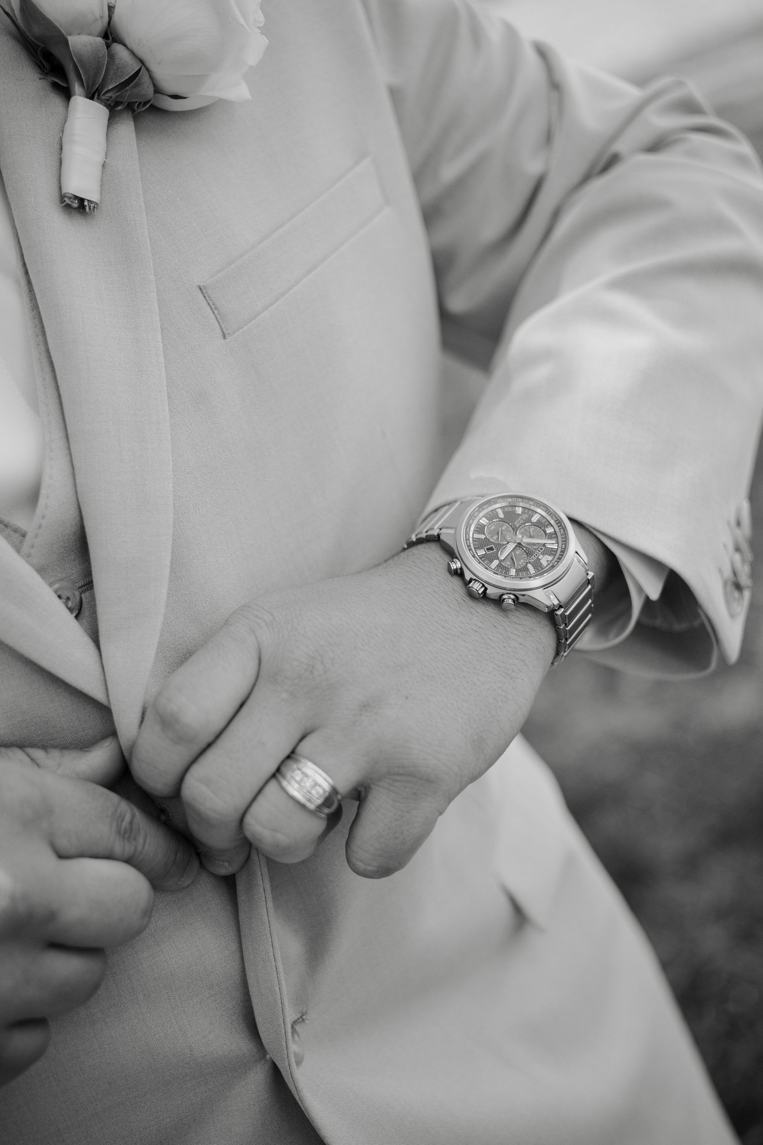 Black and white photo of groom buttoning suit jacket, wearing luxury watch and wedding ring.