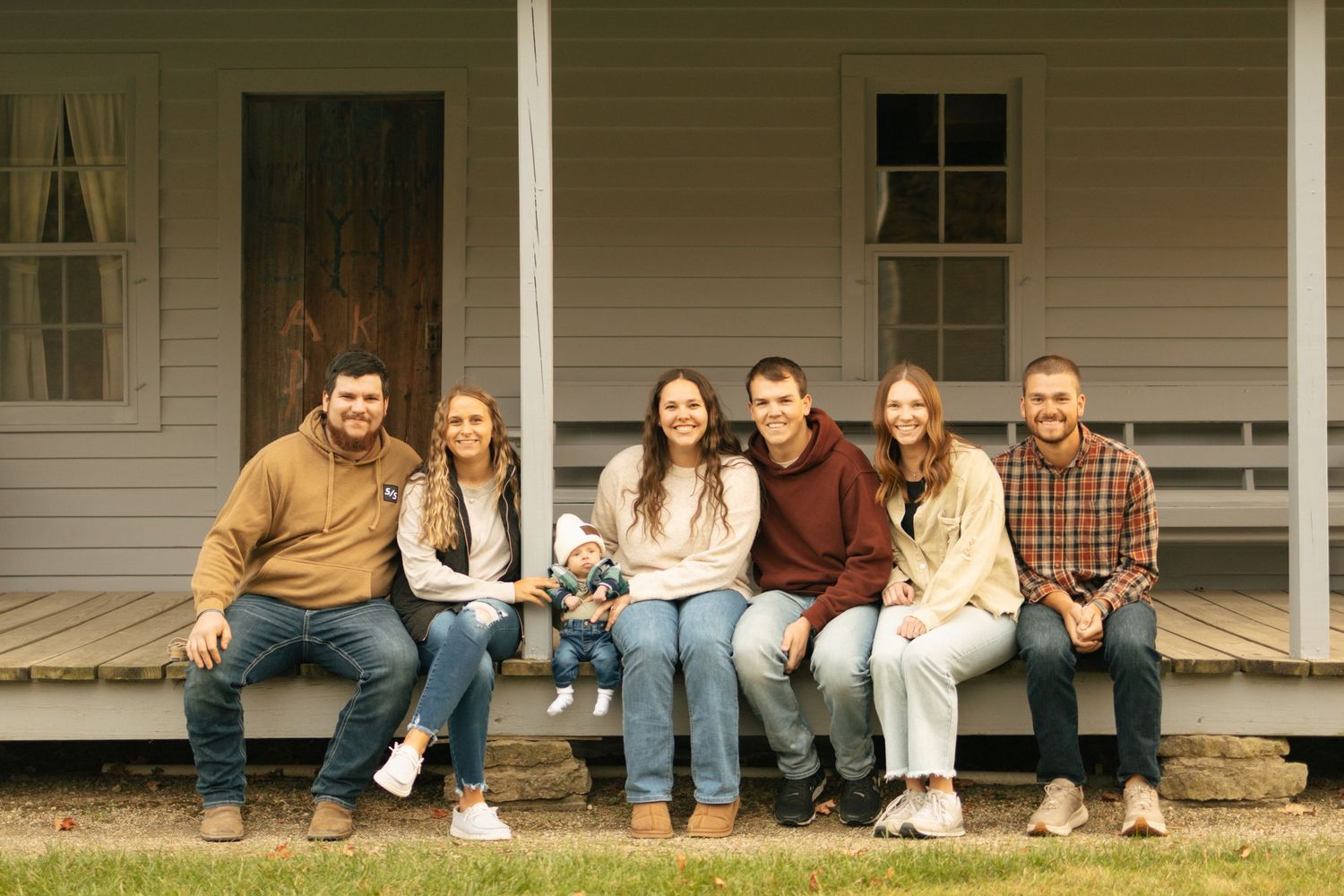 Family of six smiling on farmhouse porch, casually dressed in jeans and sweaters, with a baby in the center.