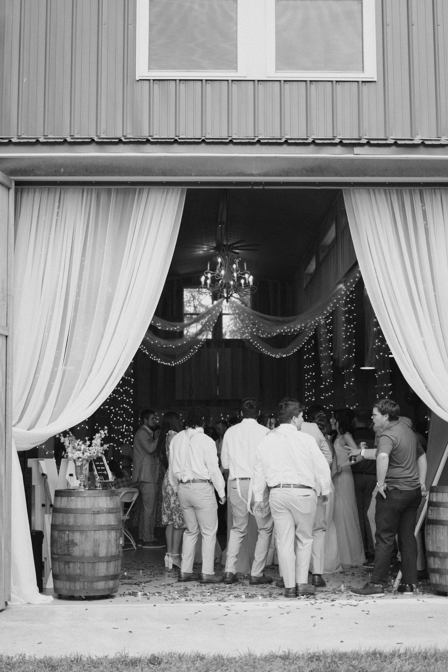 Black and white photo of wedding guests entering decorated barn venue with draping curtains and barrels.