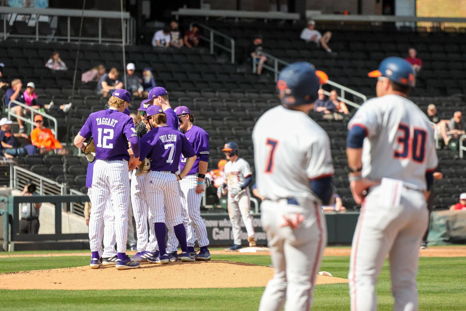 Baseball players in purple uniforms gather on the field as opposing team players in white uniforms look on during a game.