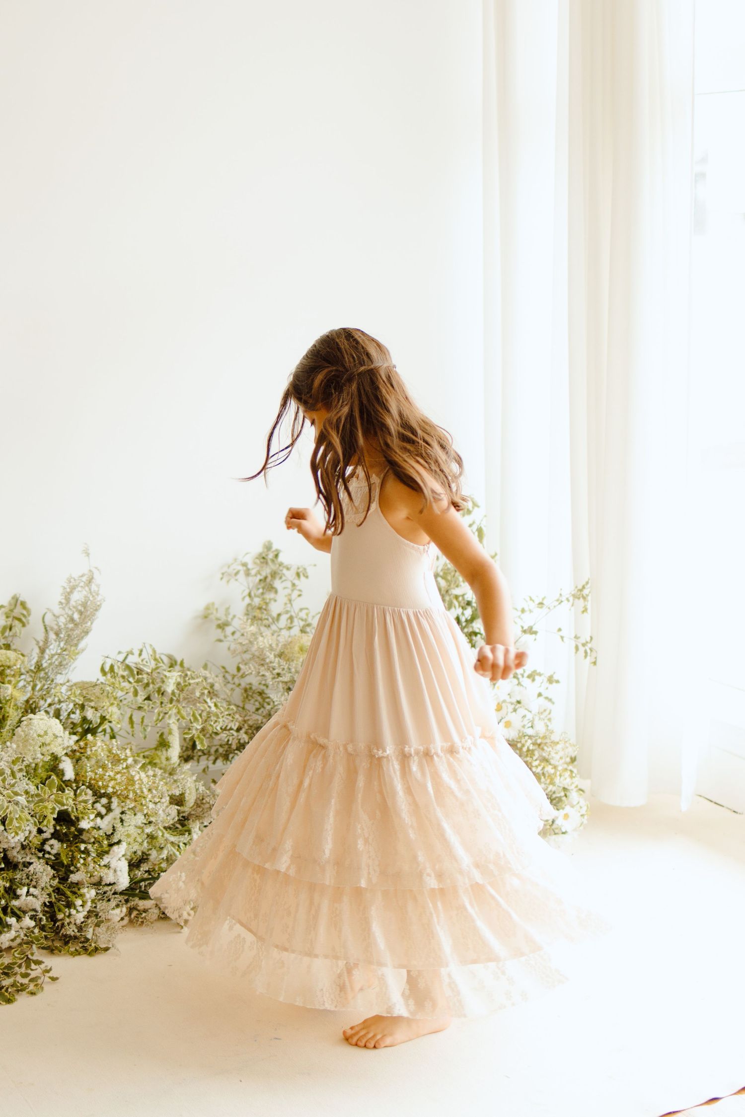 A young girl twirls in a blush tiered tulle dress surrounded by white florals in a bright studio.
