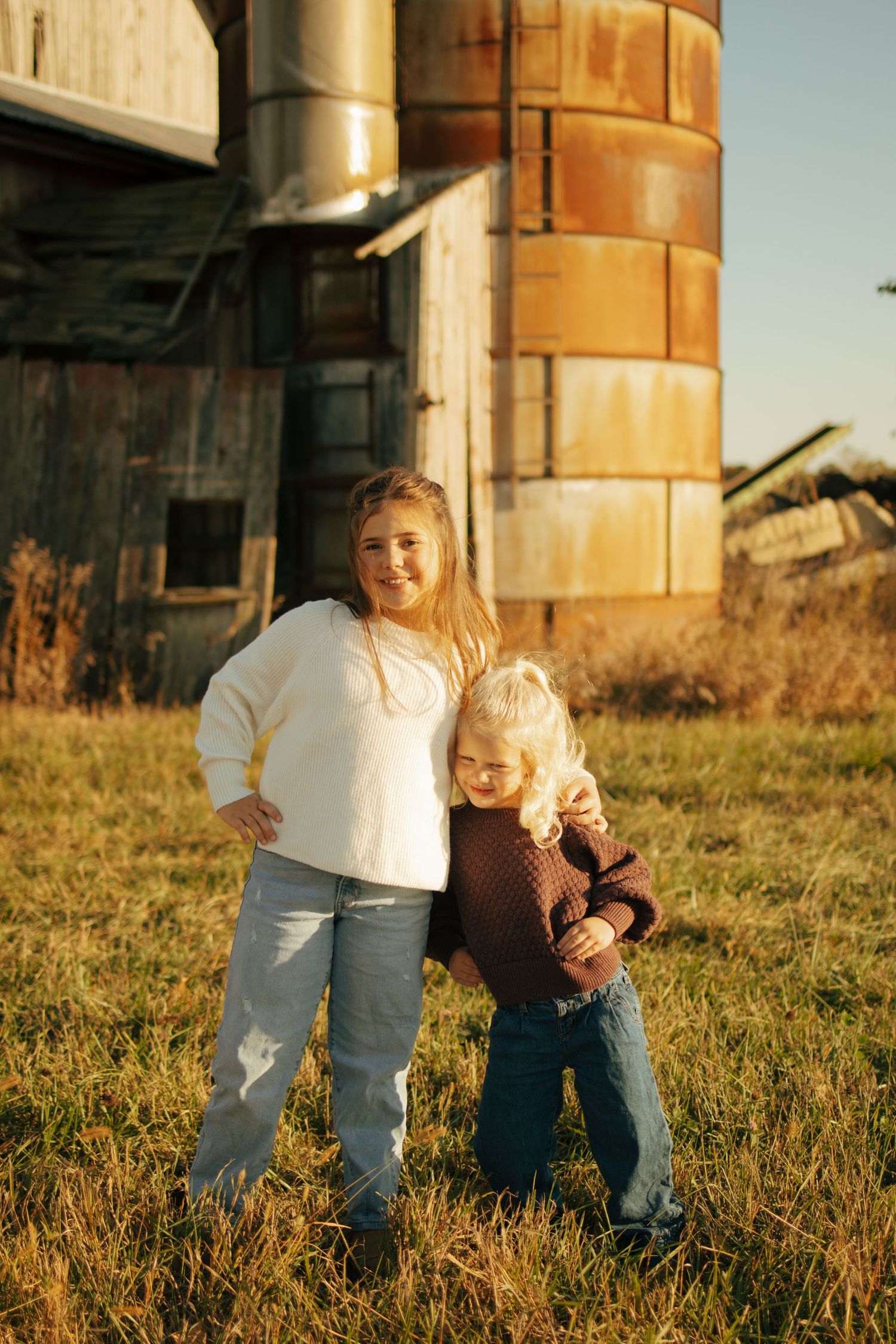 Two young blonde children pose together in a grassy field beside a rustic grain silo on a sunny day.