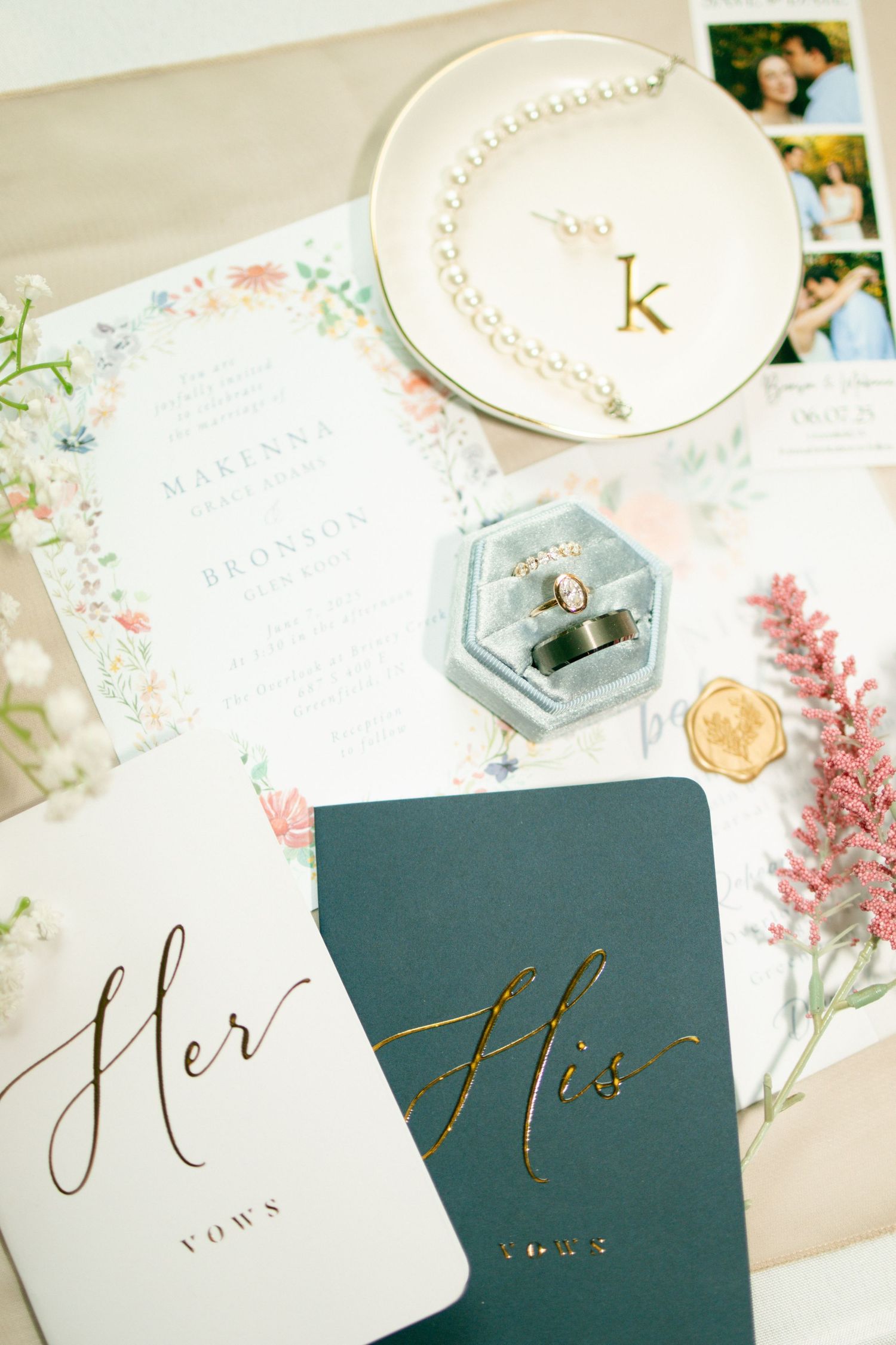 Wedding flat lay with vow books, floral invitations, pearl necklace, engagement rings in velvet box, and gold monogram dish.