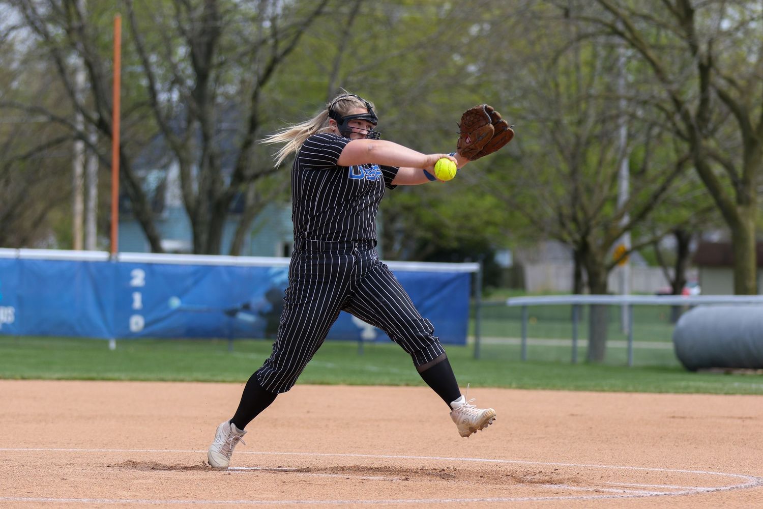 Softball player in pinstriped uniform winds up for a pitch on a dirt field during a game.