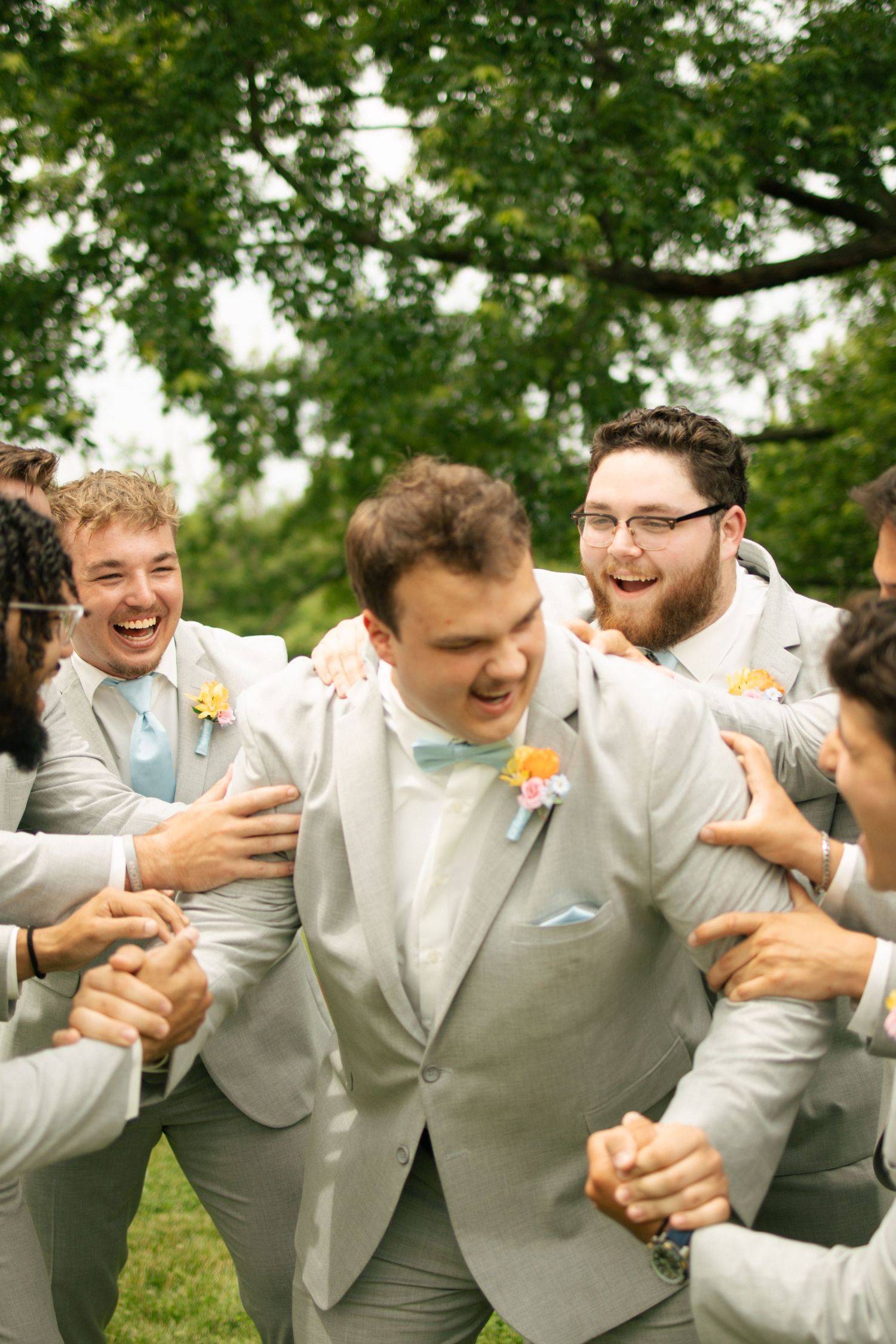 Groom laughs joyfully surrounded by groomsmen in matching light gray suits with blue bow ties outdoors.