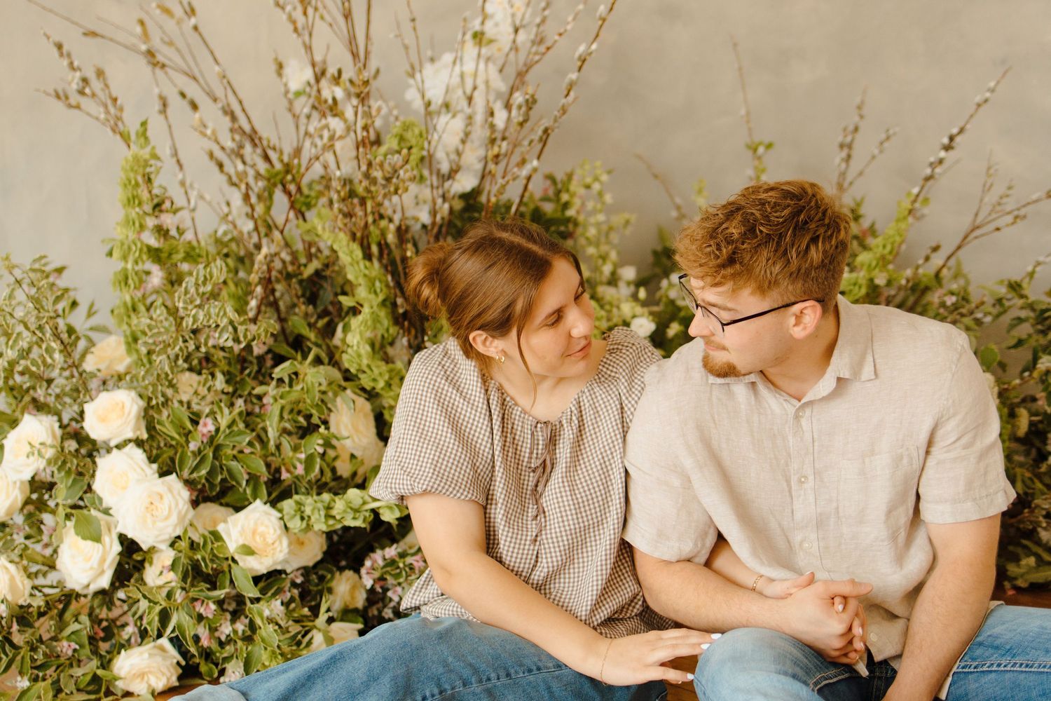 A couple sits together among lush white flowers and greenery, gazing at each other warmly in a romantic studio setting.