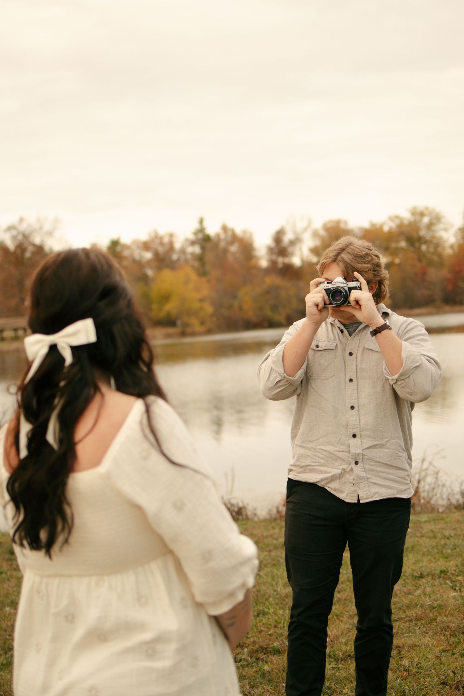 Man photographing woman with film camera near autumn lake, she wears white dress with hair bow.