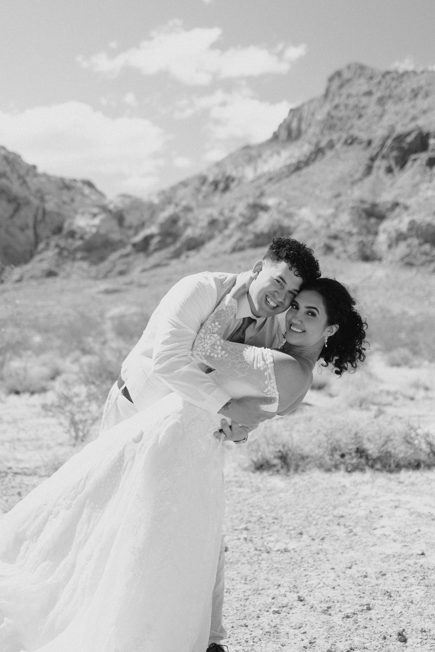 Black and white wedding photo captures newlyweds embracing joyfully against scenic desert mountain landscape.