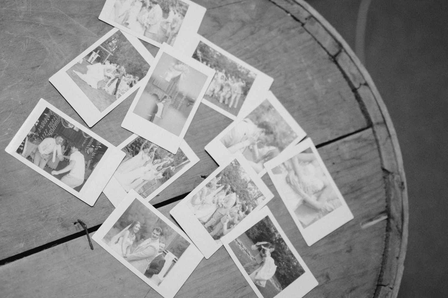 Black and white overhead view of scattered Polaroid wedding photos on wooden barrel surface.