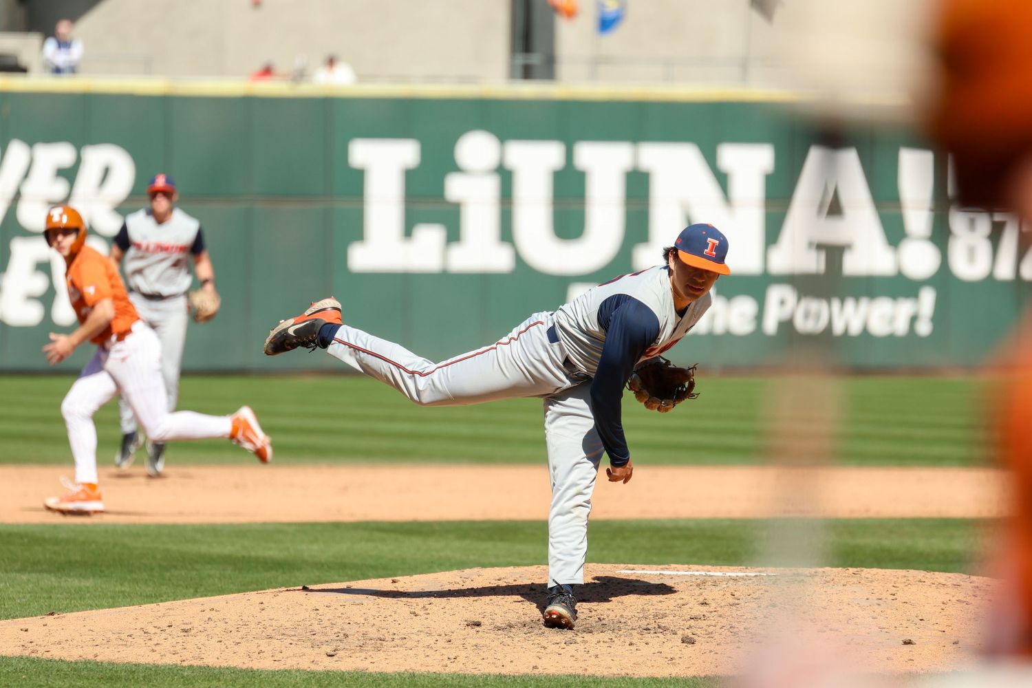 Baseball pitcher in navy uniform follows through on delivery during game with green outfield wall in background.