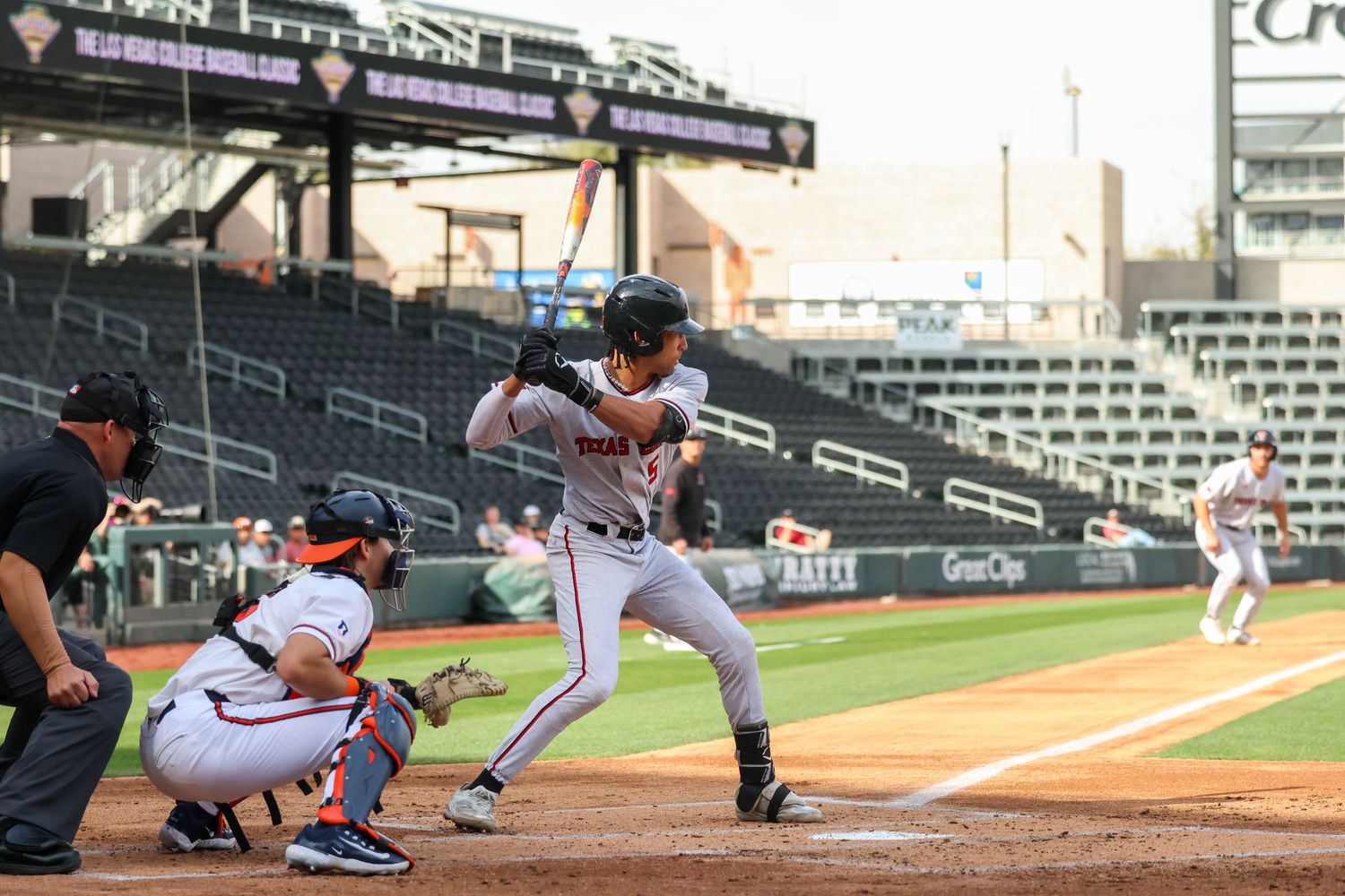 Baseball batter in grey uniform swings at pitch during game with empty stadium seats visible in background.