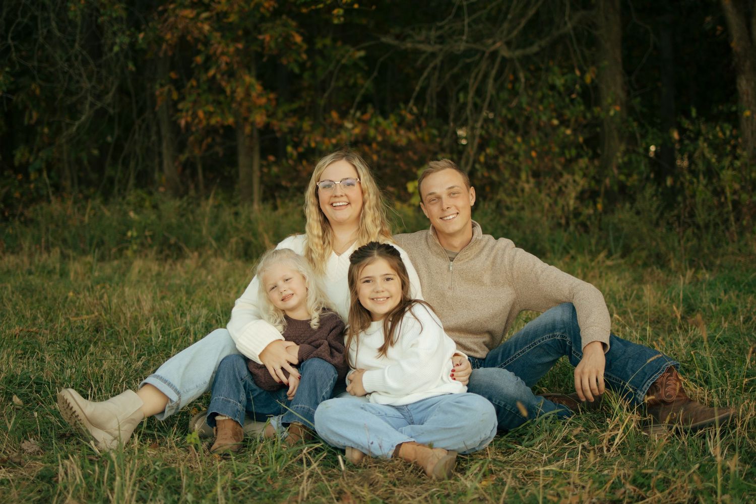 Family of four sitting together on grass in front of autumn trees, smiling warmly in a fall outdoor portrait.