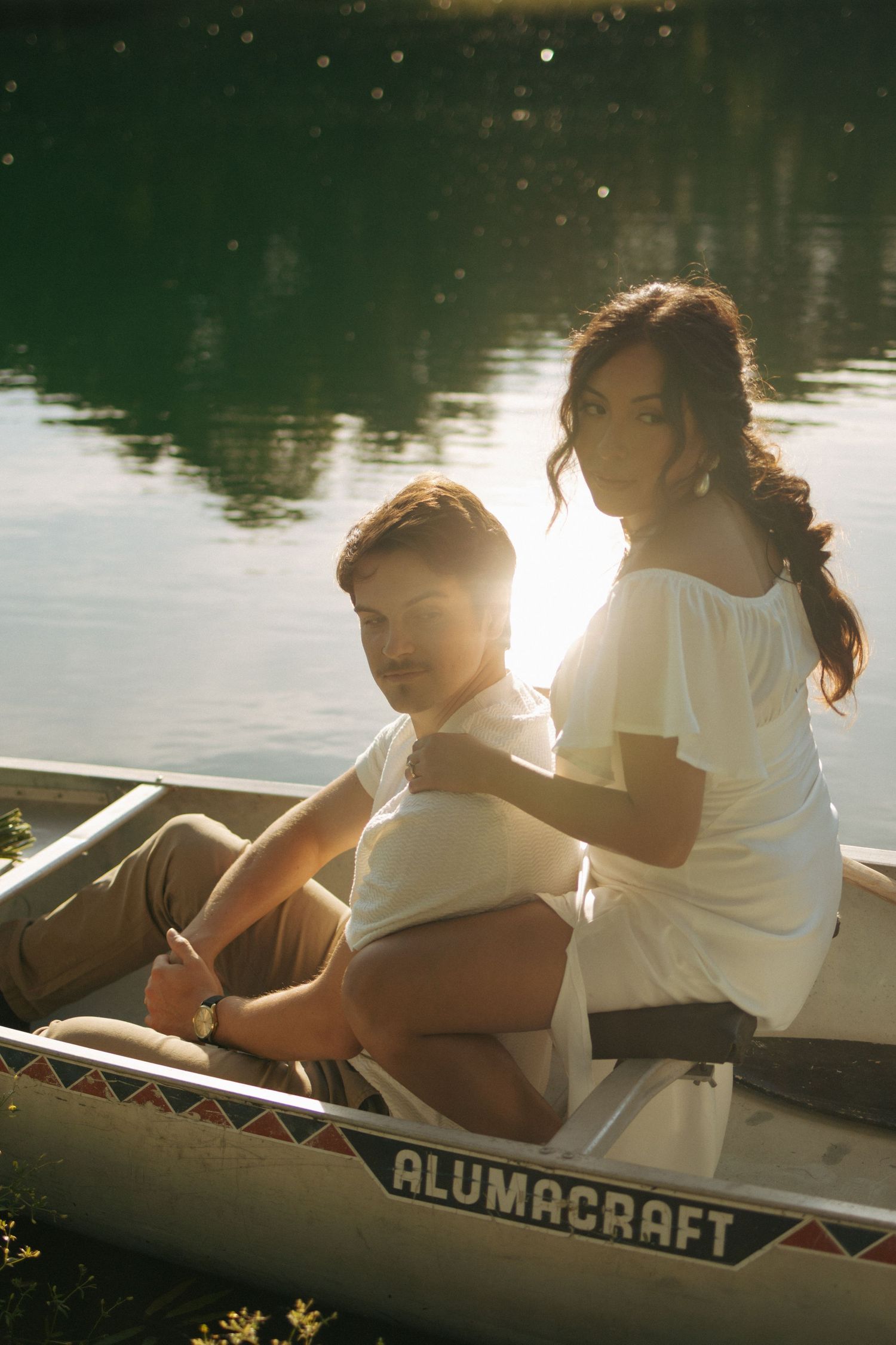 Couple sitting together in an Alumacraft canoe on a sunlit lake, woman in white dress embracing man from behind.