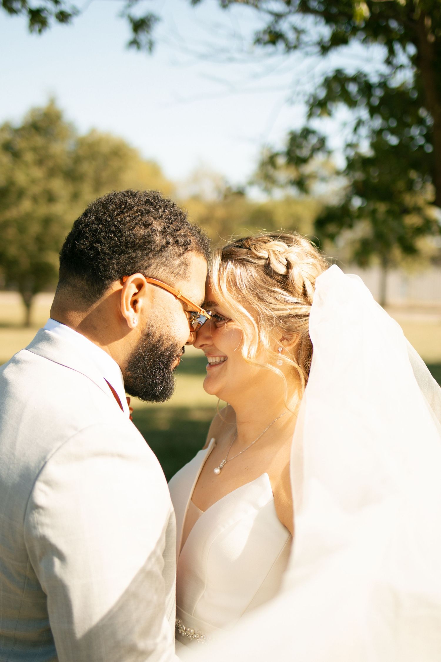 Bride and groom touching foreheads romantically outdoors in golden sunlight, bride wearing veil and groom in white suit.