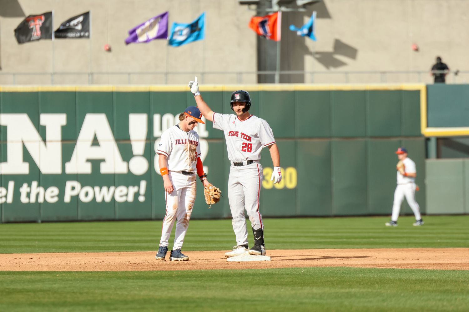 Two baseball players stand together on a baseball field during a game with a green wall in the background.