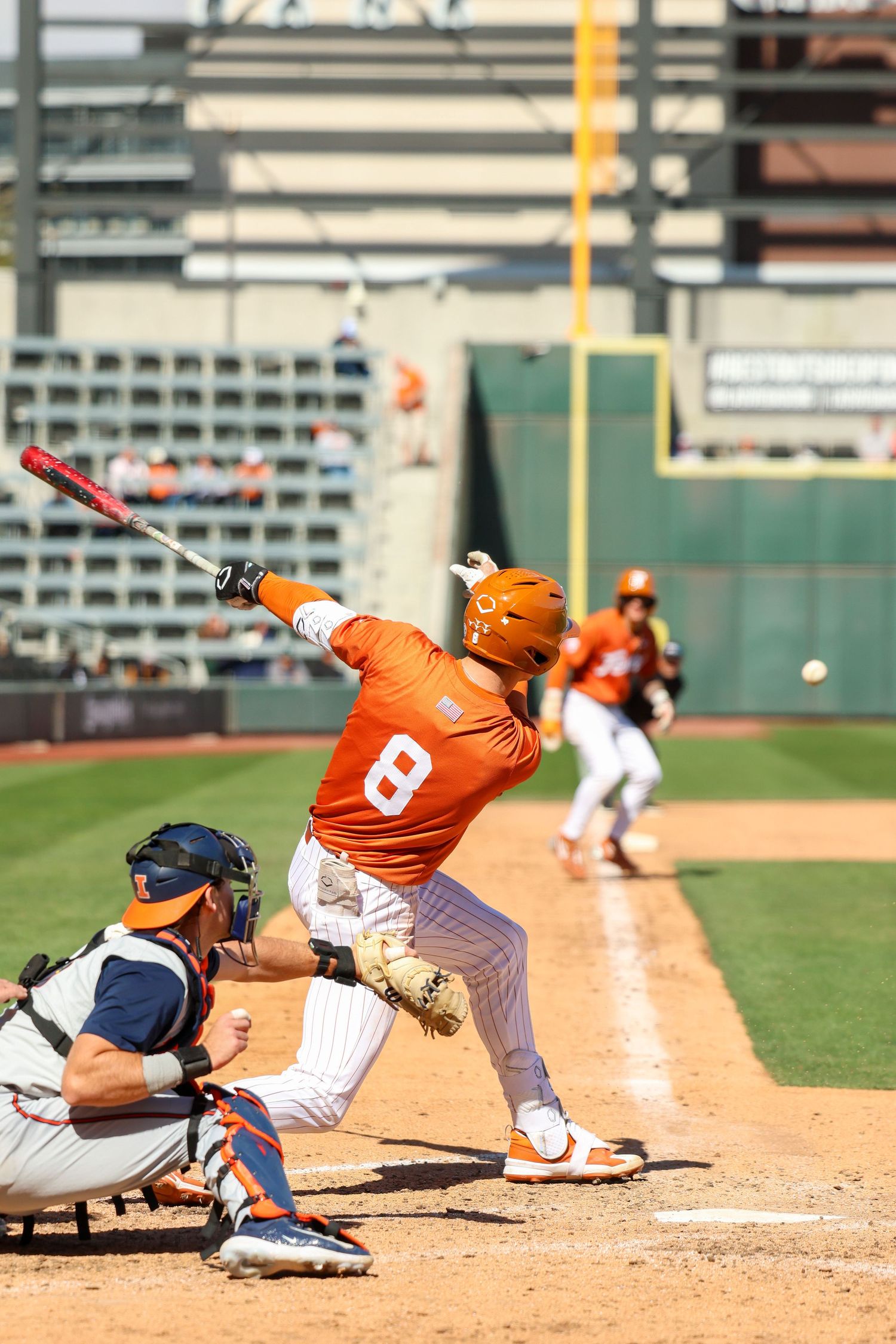 Baseball player in orange uniform swings bat at home plate during game with catcher in navy crouched behind plate.