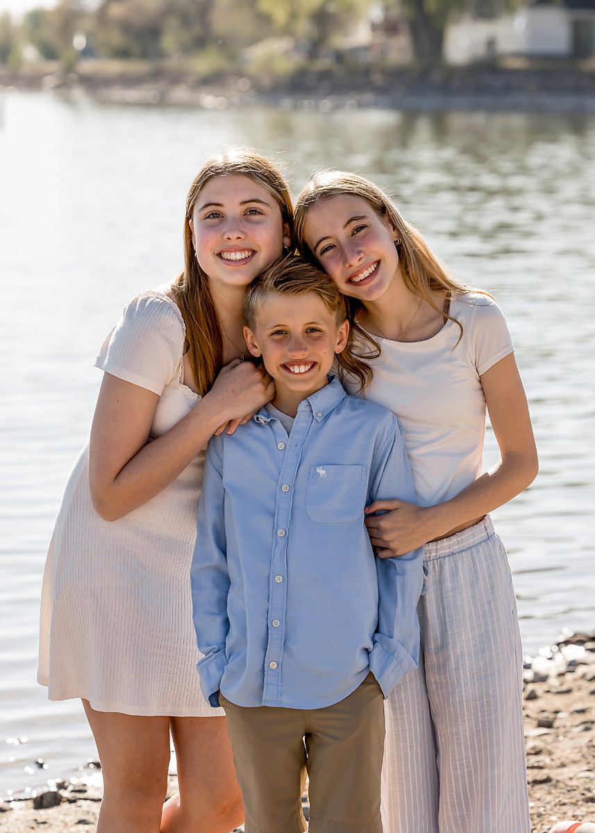 Two teenage girls and a young boy pose together smiling by a lake.