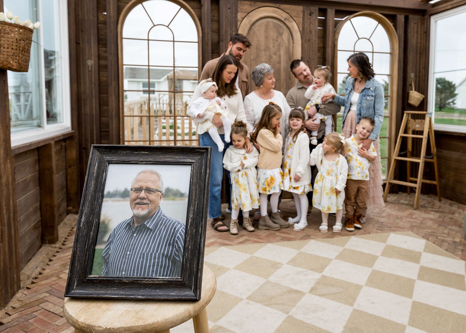 Family gathered around a framed memorial portrait of an elderly man in a warm rustic greenhouse setting.