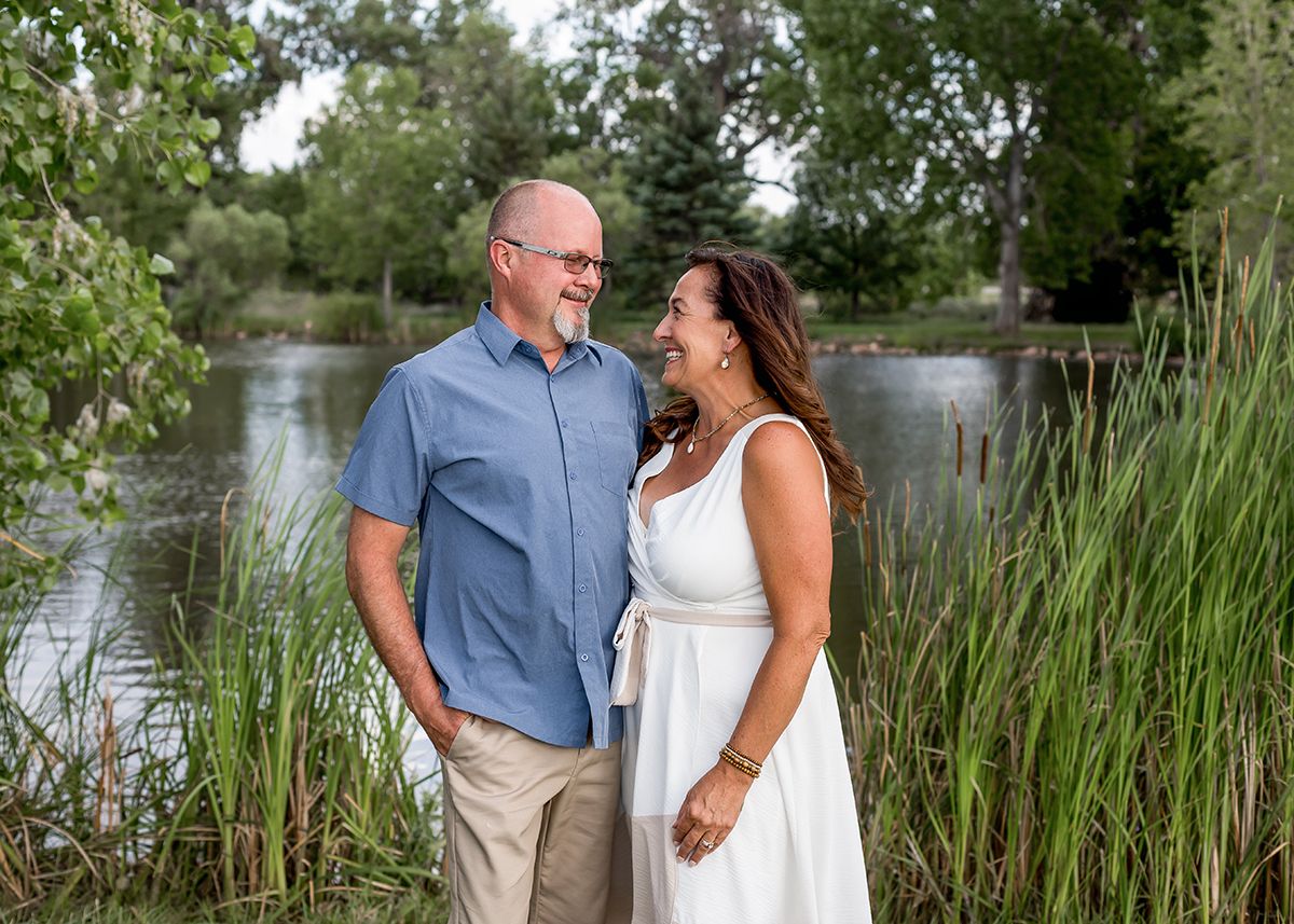 Couple smiling at each other near a pond surrounded by tall grass and trees on a sunny day.