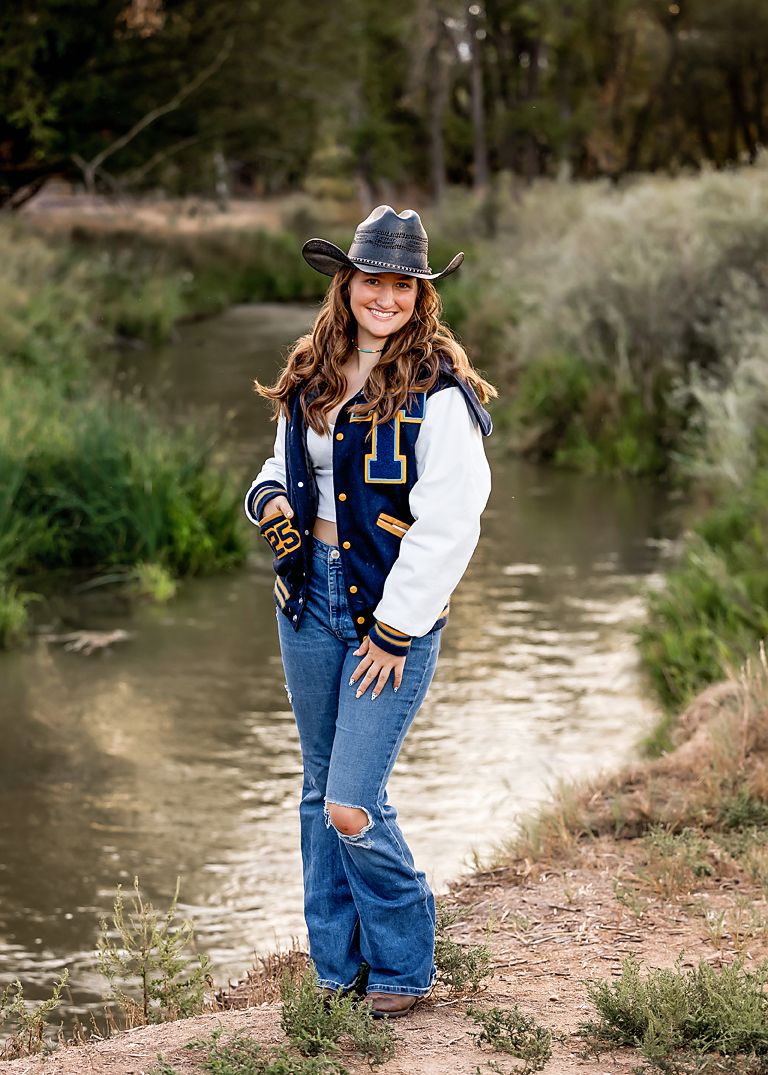 Young woman in varsity jacket, cowboy hat, and ripped jeans poses confidently by a serene creek surrounded by lush greenery.