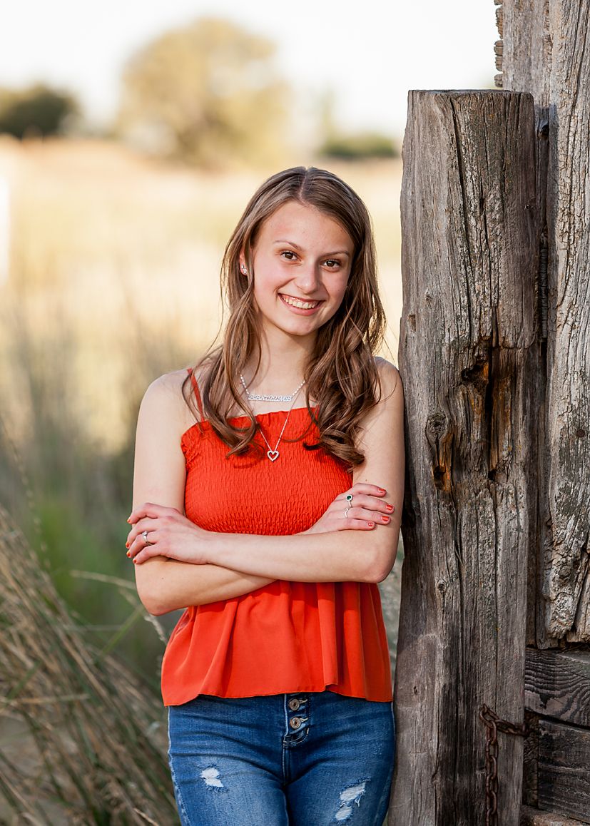 Teen girl in red top and jeans leans against rustic wooden post in sunny field.