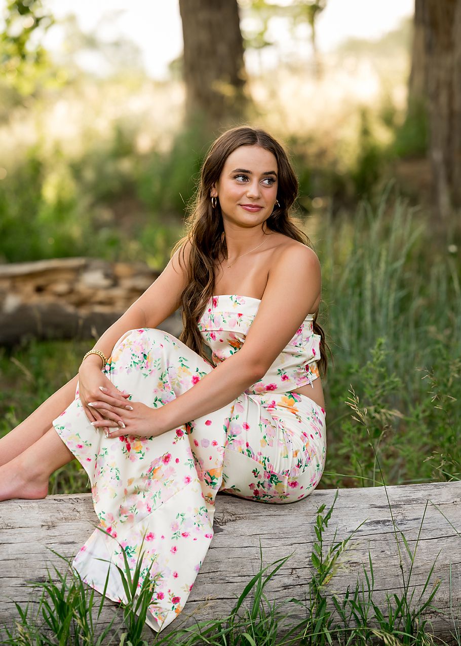 Teen girl in floral jumpsuit sits on log in lush green outdoor natural setting.