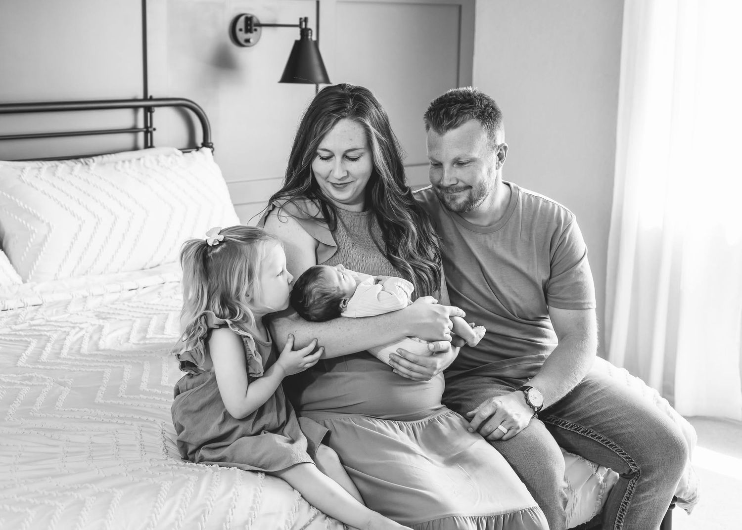 Black and white family photo with parents and daughter admiring newborn on bed.