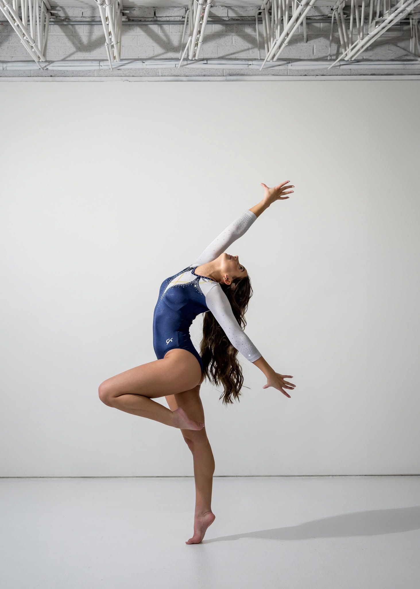 Gymnast in navy and white leotard performs elegant back arch pose balanced on one leg in studio.