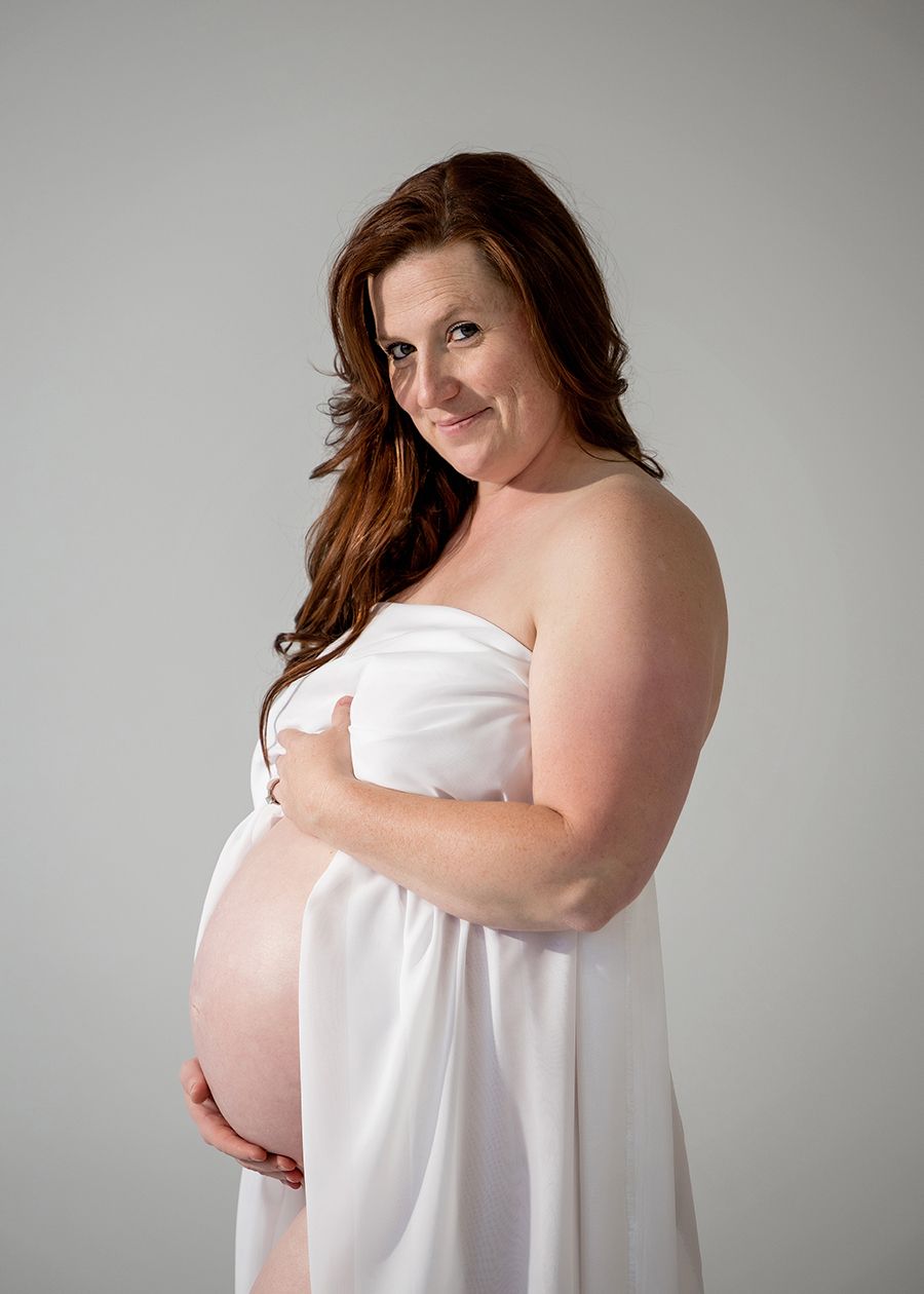 Pregnant woman in white strapless dress cradling baby bump, smiling softly at camera in professional maternity photo studio.