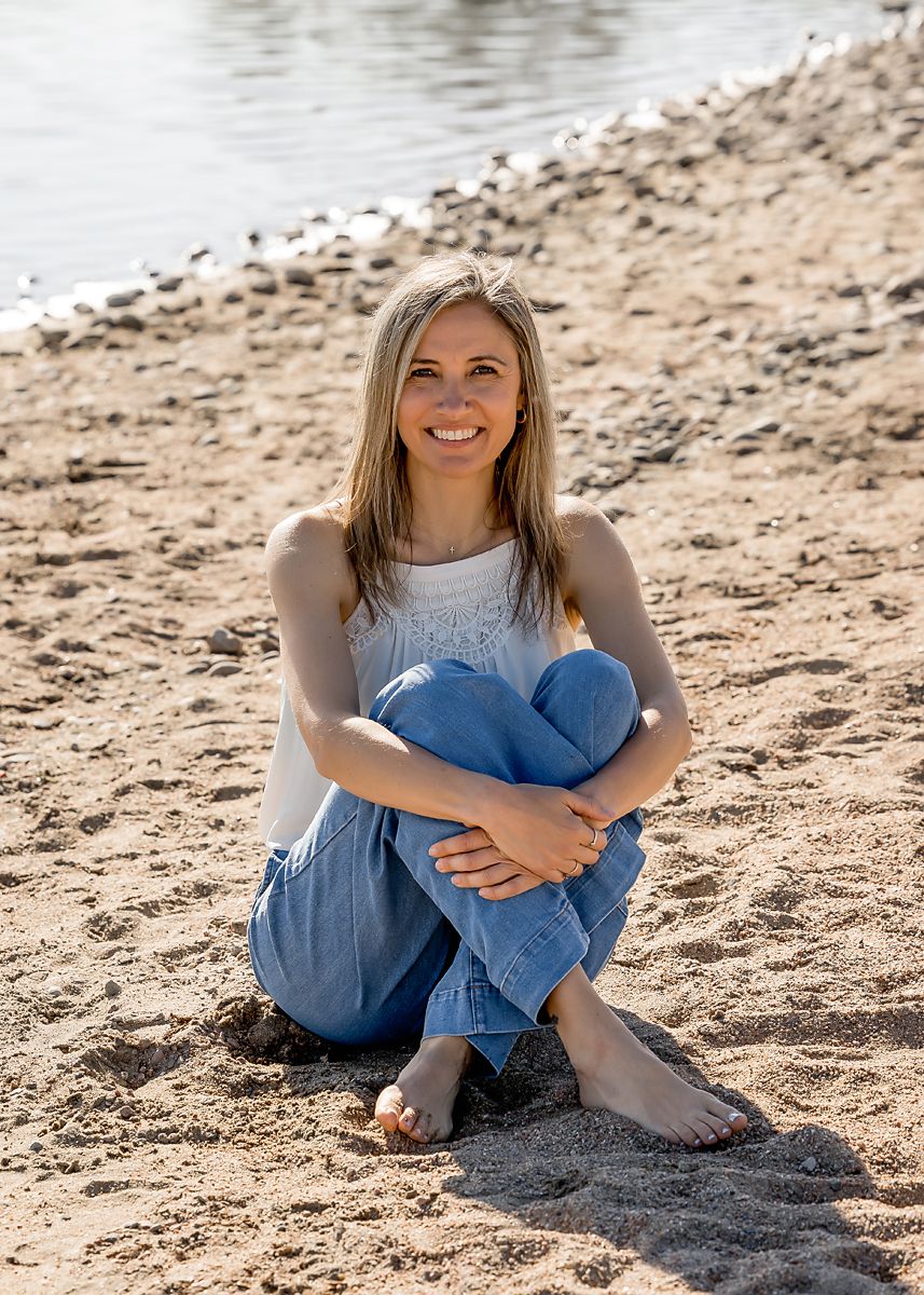 Smiling blonde woman sitting barefoot on sandy beach in white top and blue jeans near water.