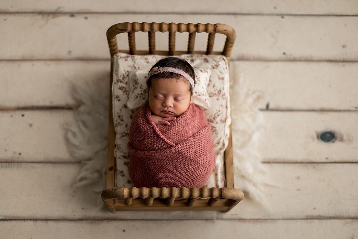 Newborn baby girl swaddled in pink wrap sleeping in a tiny wooden bed with floral bedding.