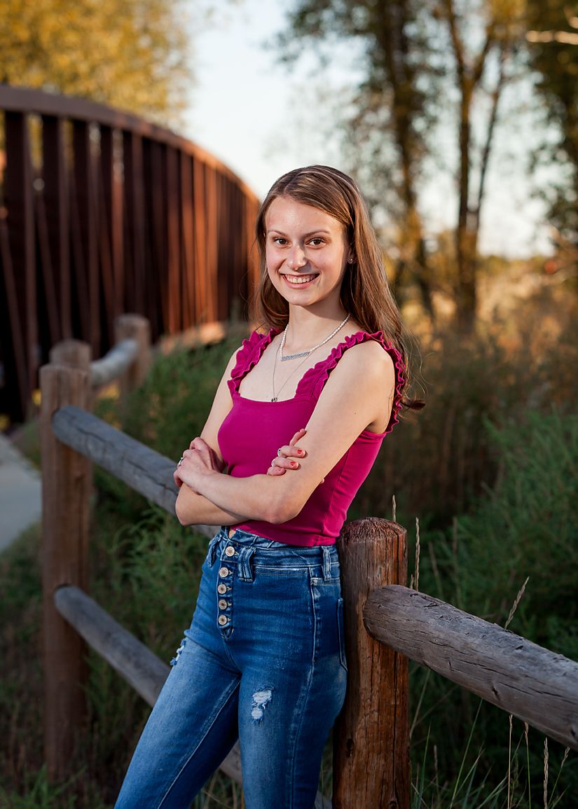 Smiling brunette teen in pink tank top and jeans leans against a wooden fence near a bridge outdoors.